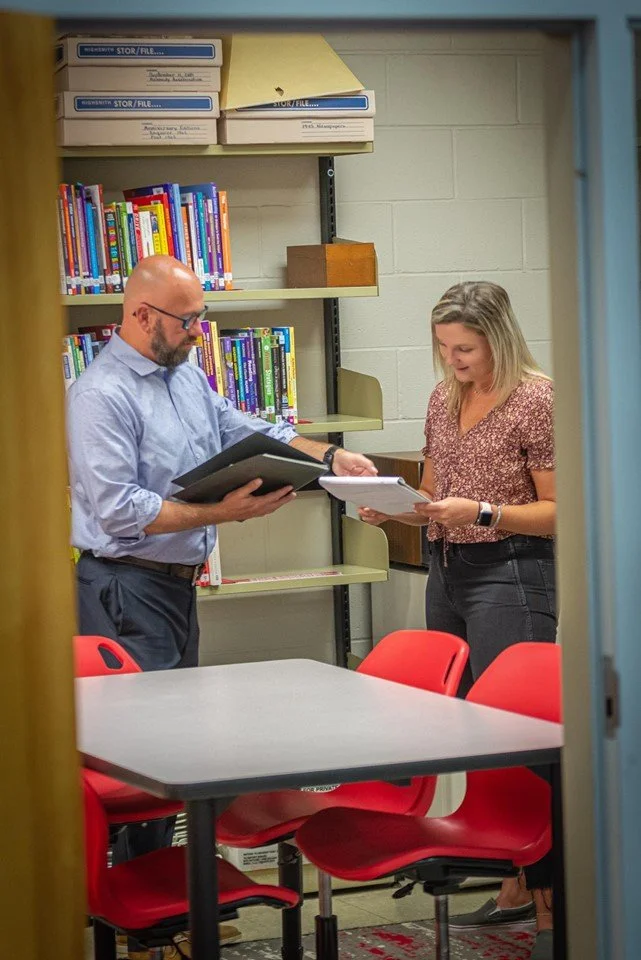 A man and woman look at documents and folders in a room with a bookshelf, some red chairs, and a table.
