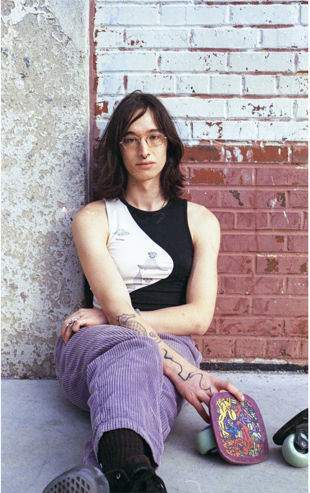 Young woman with glasses sitting against a textured beige and red brick wall, wearing a sleeveless black and white top, purple corduroy pants, and black sneakers, holding a colorful skateboard.