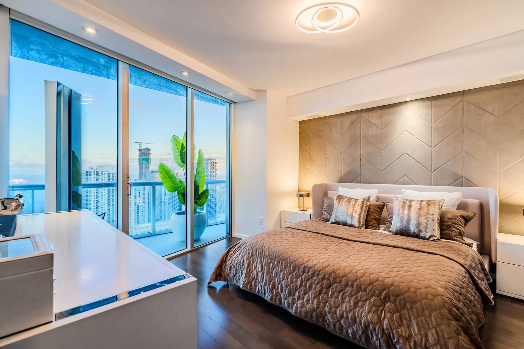 Modern bedroom with a large bed, decorative pillows, a nightstand with a lamp, a textured accent wall, and a balcony with floor-to-ceiling windows showing a city skyline.