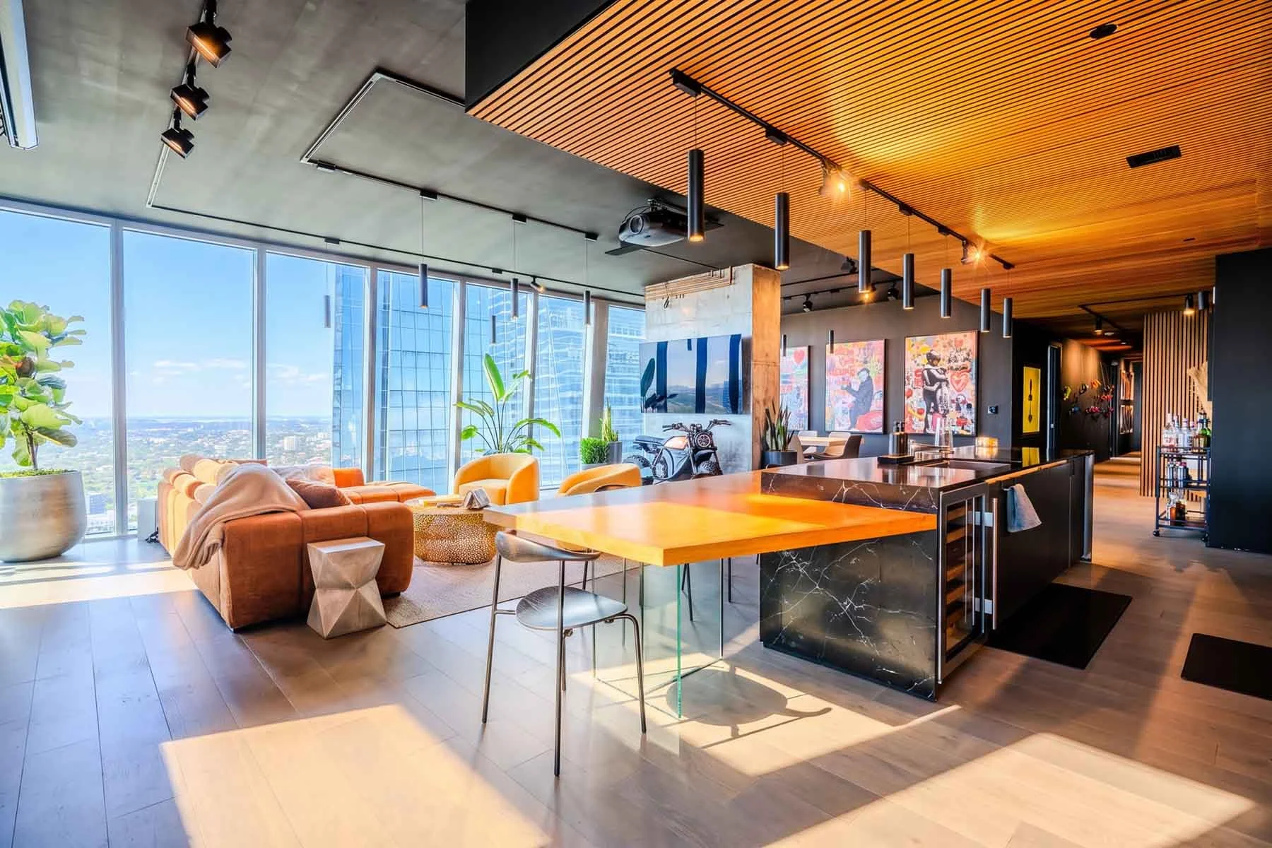 Modern high-rise apartment living room with large windows, tan sofa with blankets, yellow armchair, wooden dining table, black marble kitchen island, and colorful artwork on the walls.