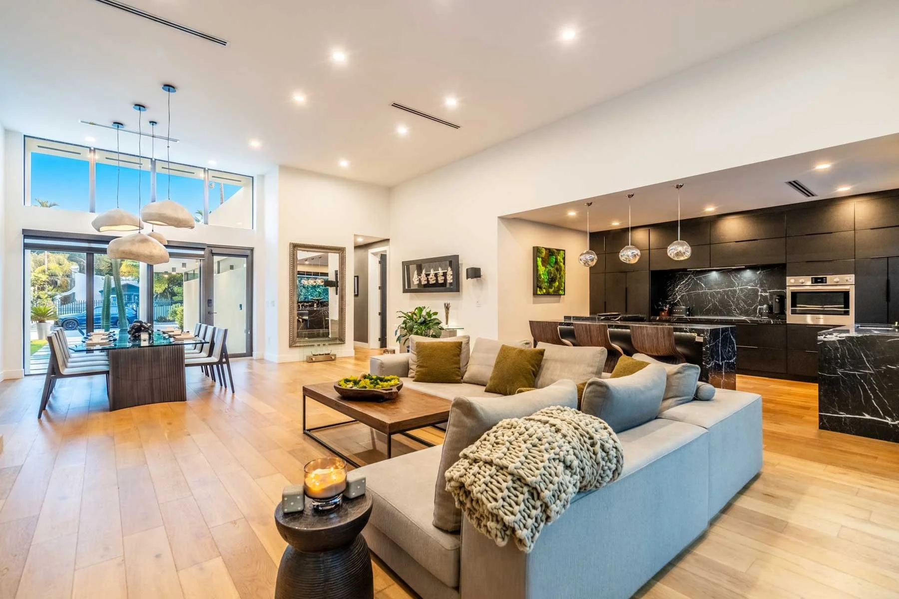 Open-concept living room and dining area in a modern home with large windows, a black marble kitchen island with pendant lights, a beige sofa with cushions, a wooden dining table with chairs, hardwood floors, and decorative plants.