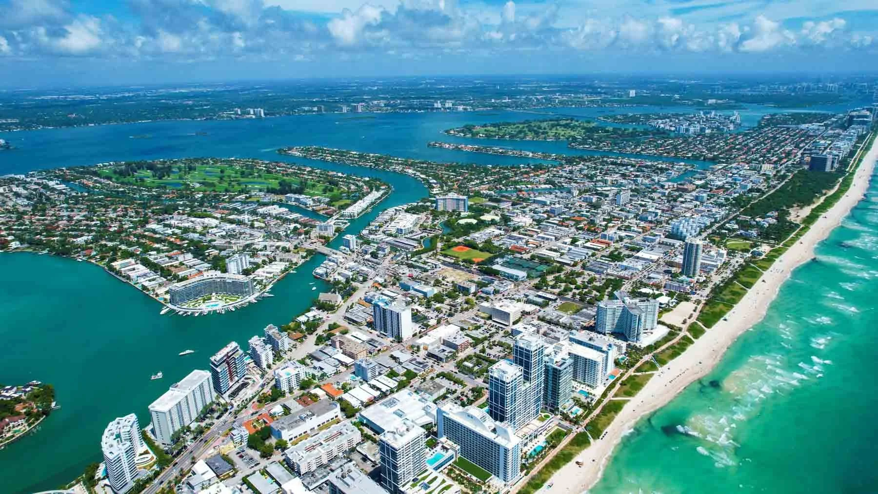 Aerial view of a coastal city with a beach on the right side, high-rise buildings, and a river flowing through the city, with a mix of urban areas and green parks.