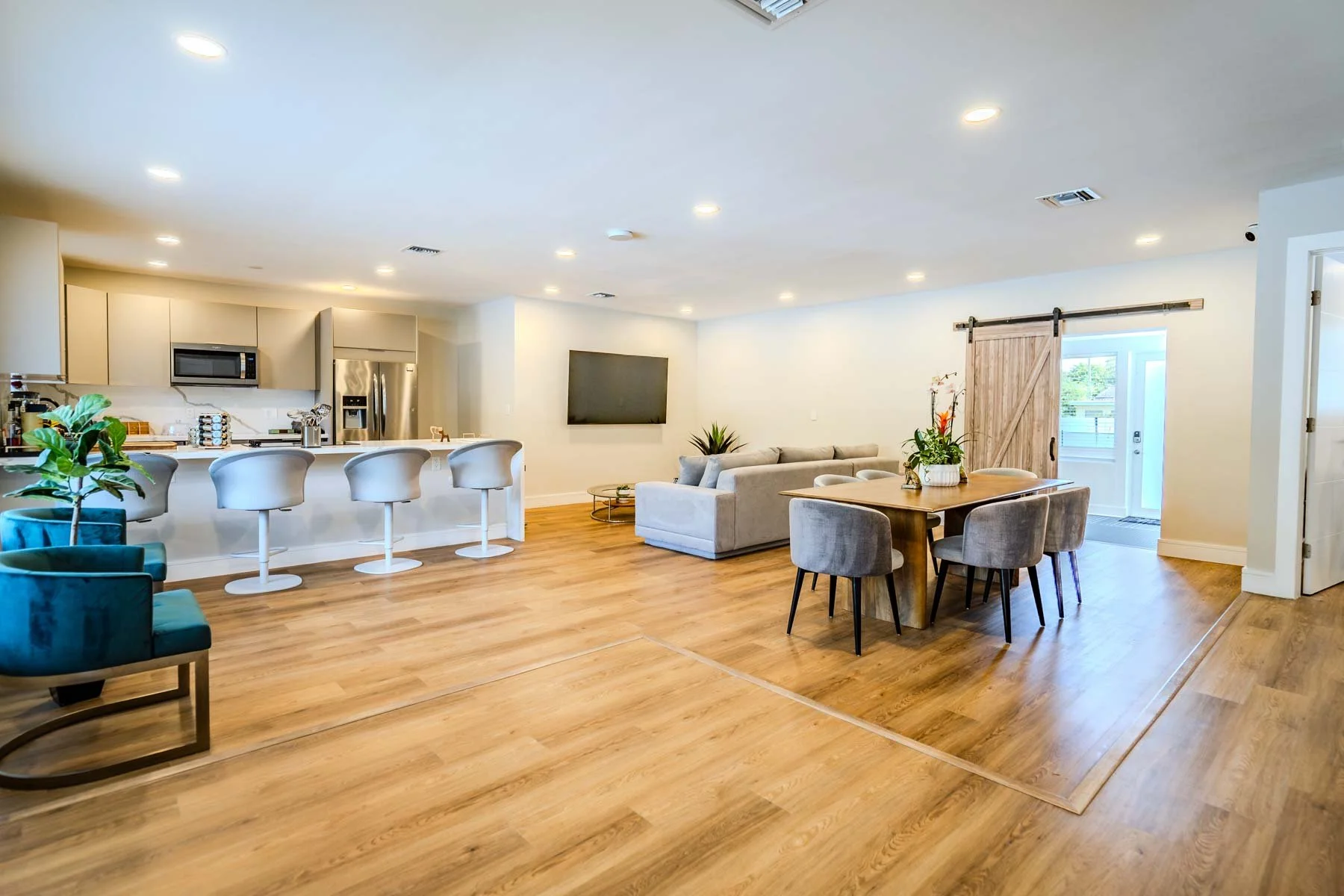 An open-concept living space with a kitchen, dining, and seating area. The kitchen has a modern design with white cabinets, a stainless steel refrigerator, microwave, and a white island with four barstools. The living area has a wall-mounted TV, a li