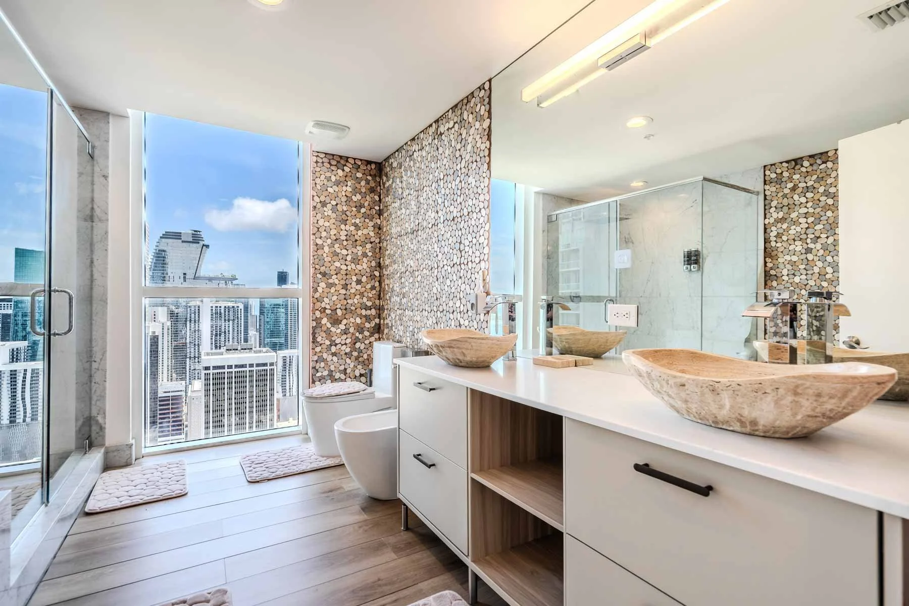 Modern bathroom with double vessel sinks made of stone on white countertop, large mirror, and city skyline view through floor-to-ceiling window, wooden flooring, small white rugs, and a glass shower enclosure.