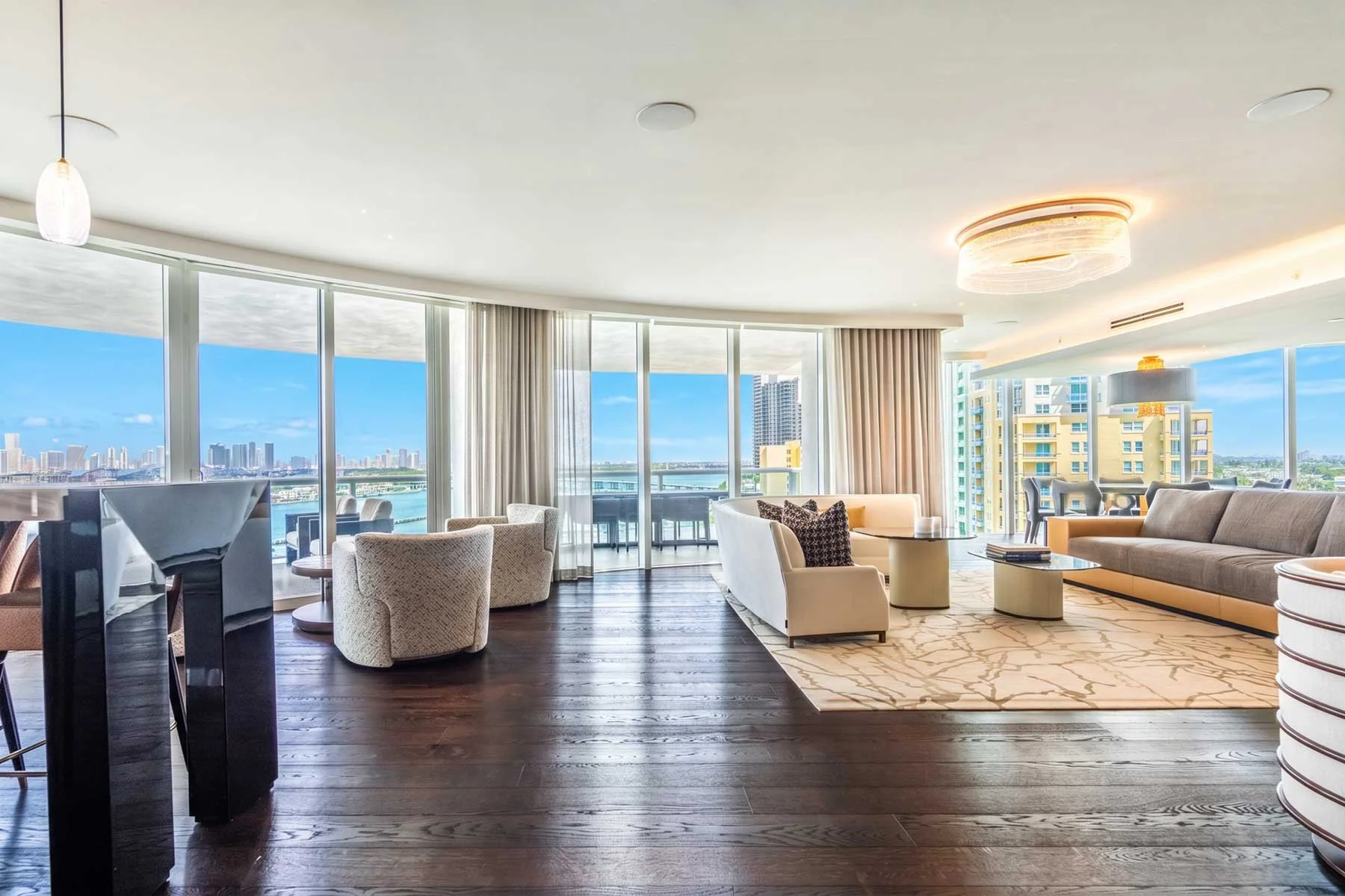 Living room with large floor-to-ceiling windows showing a city skyline and water view, modern furniture including sofas, armchairs, a coffee table, and a dining table with chairs, hardwood floors, and ceiling lights.