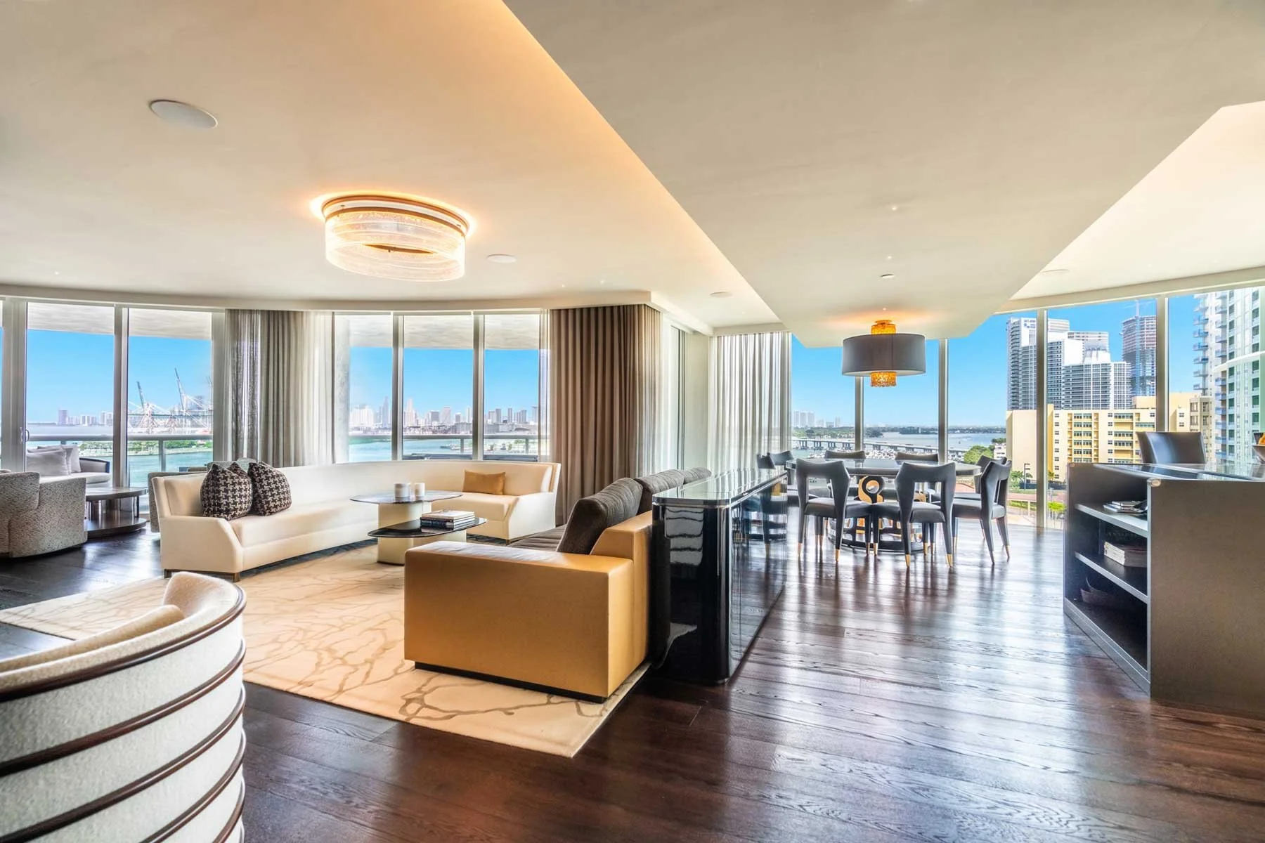Modern living room with large windows showcasing city and river views, featuring cream and tan furniture, a dining table with chairs, and contemporary lighting.