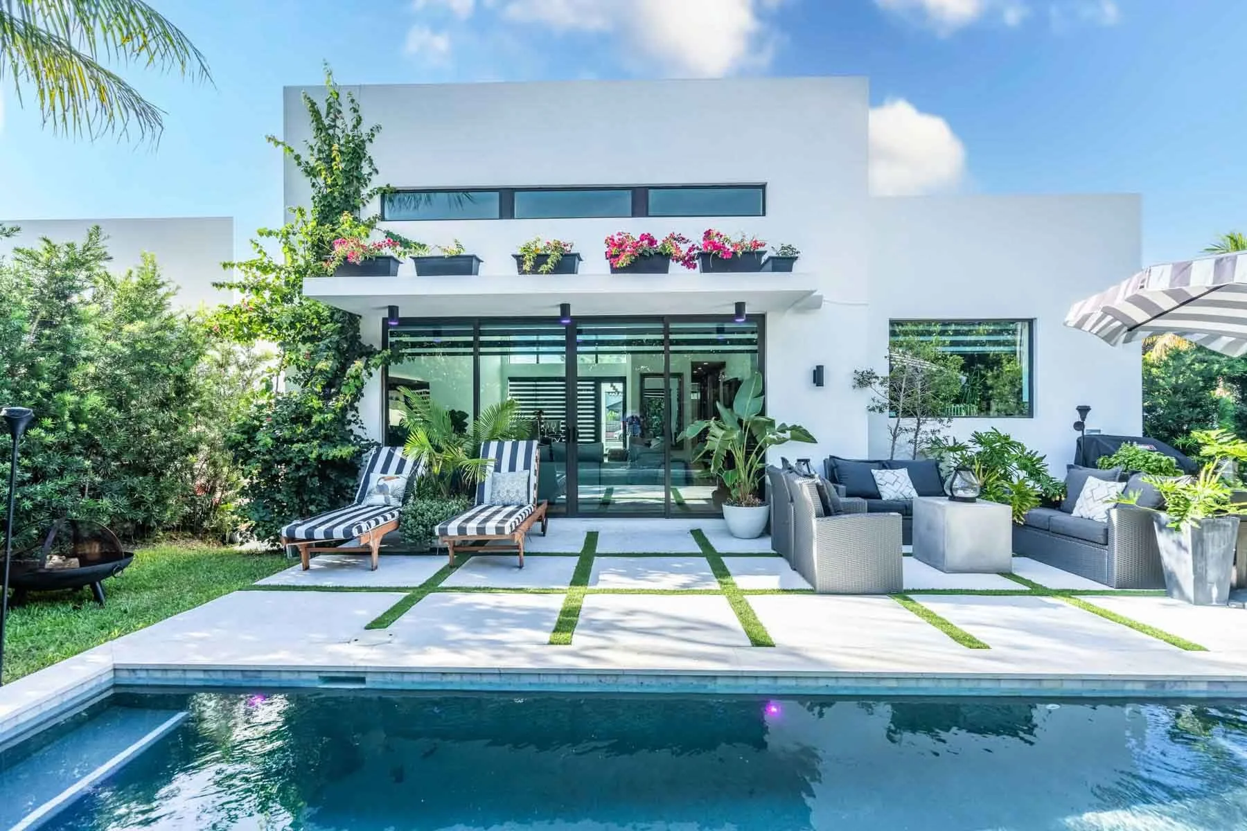 Modern white house with outdoor patio, potted plants, striped deck chairs, outdoor seating, and a swimming pool in the foreground under a partly cloudy sky.