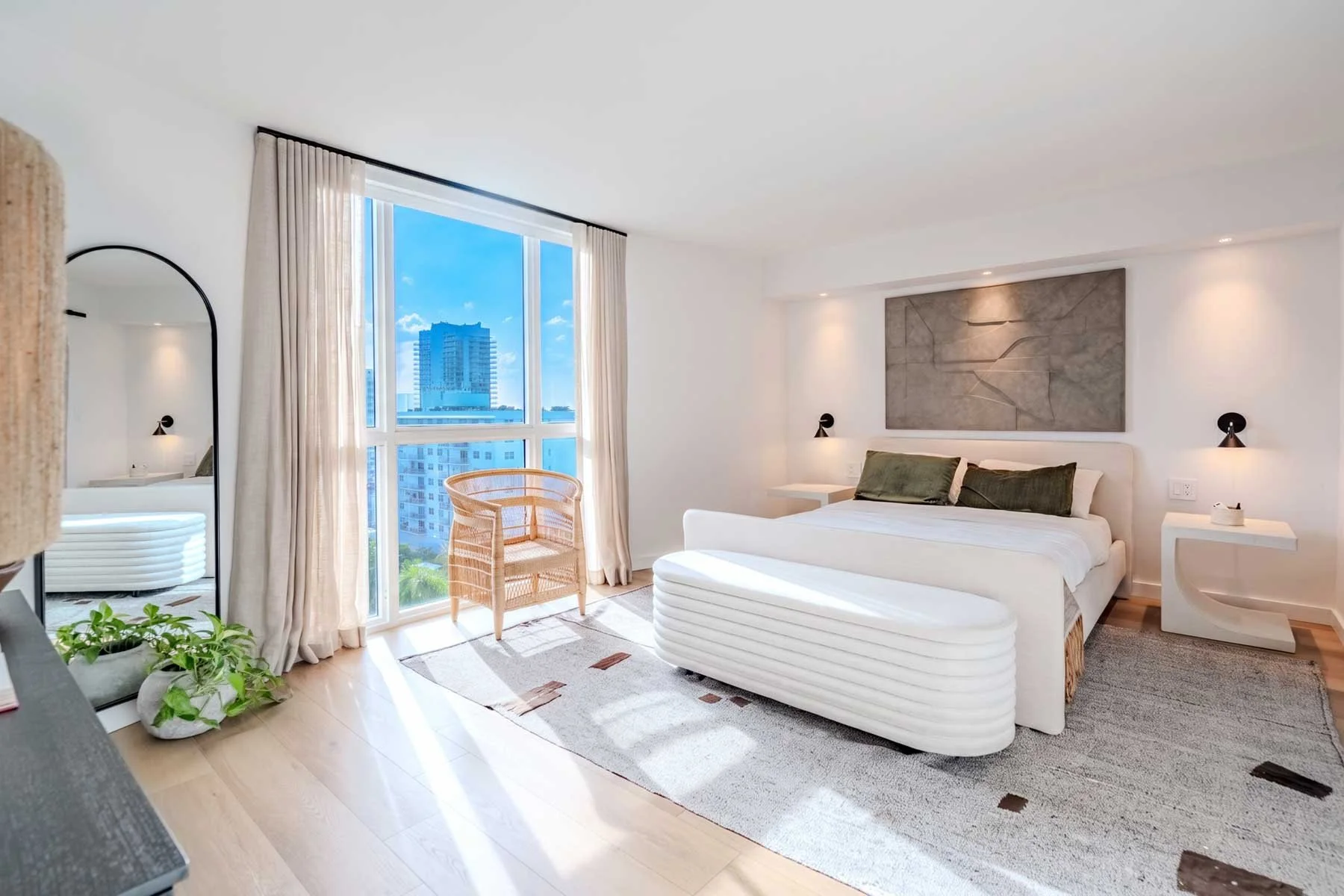 Bright bedroom with large window overlooking city skyline, white bed with dark green pillows, modern artwork above headboard, minimalist bedside tables with lamps, rattan chair by window, mirror reflecting part of the room, and a potted plant in corner.