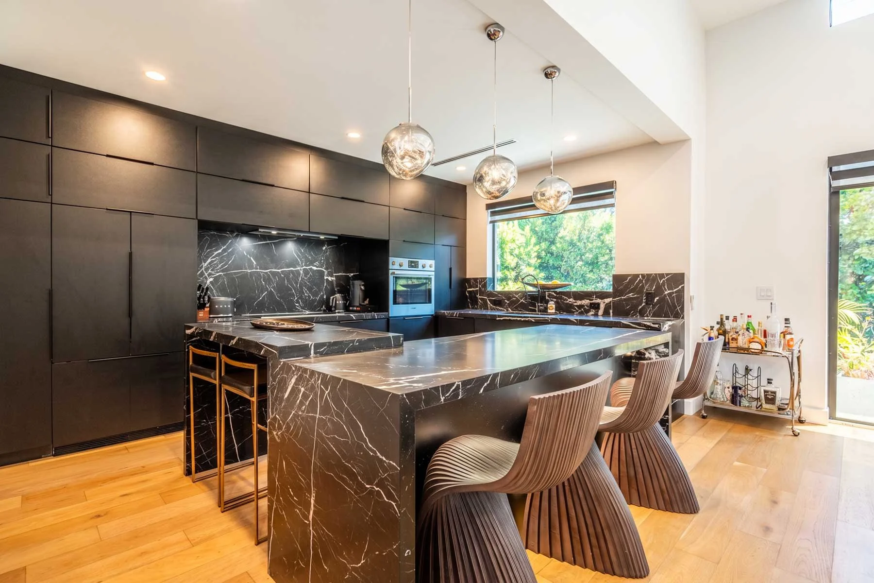 Modern kitchen with black marble countertops and backsplash, dark cabinets, wooden chairs around a marble island, and pendant lights hanging from the ceiling.