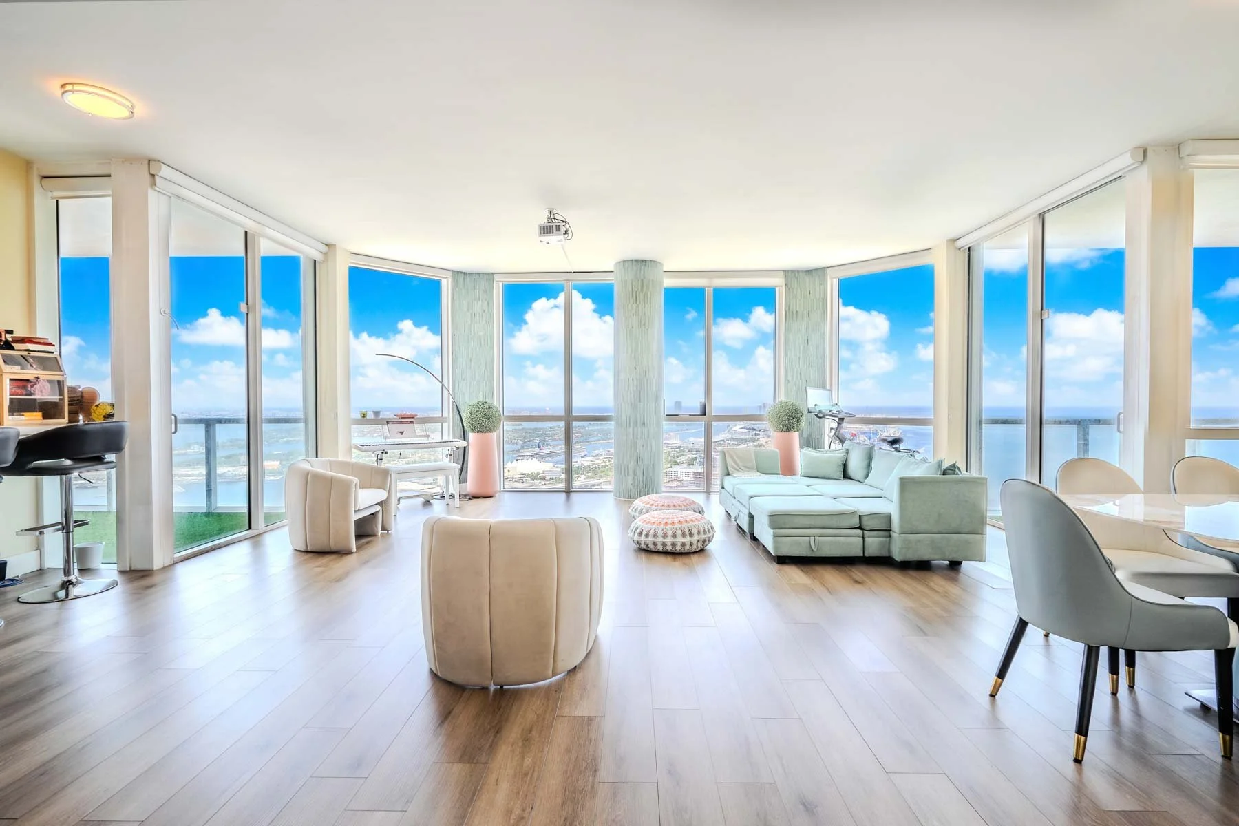 Living room with large windows showing a city view, a light-colored sectional sofa, chairs, round ottomans, plants, and a dining area.