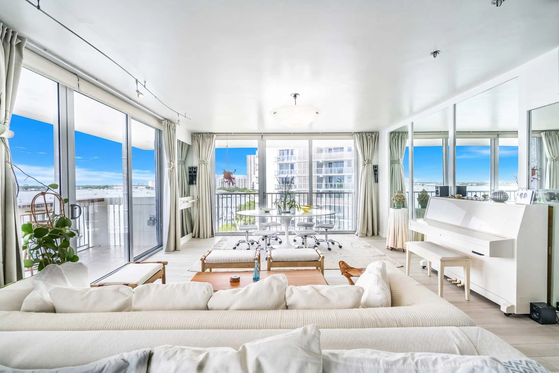 Bright living room with large glass sliding doors and windows overlooking a city skyline and water. Features white furniture, potted plants, a white upright piano, and a round dining table with chairs. Decor includes curtains, vases, and decorative i
