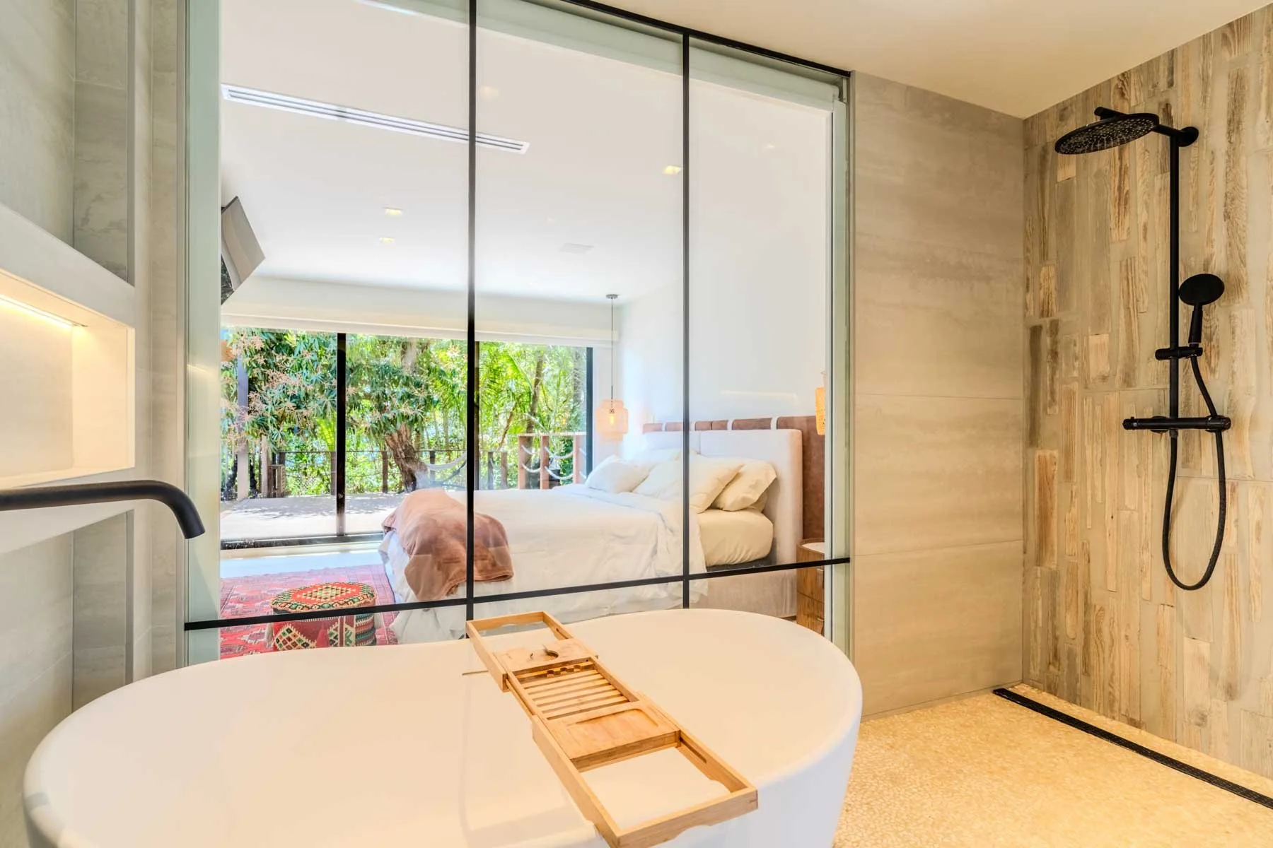 Miami Real Estate Photography. Modern bathroom with a glass partition, a freestanding bathtub, and a shower area with a rainfall showerhead. The bathroom opens to a bedroom with large windows and outdoor greenery.