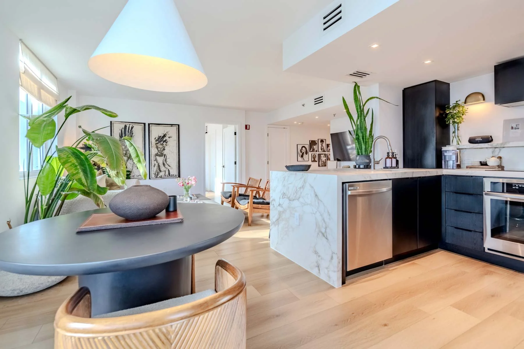 Modern open-concept kitchen and living area with white and black cabinetry, marble countertops, and wooden flooring. Decor includes potted plants, framed artwork, and a round dining table with chairs.