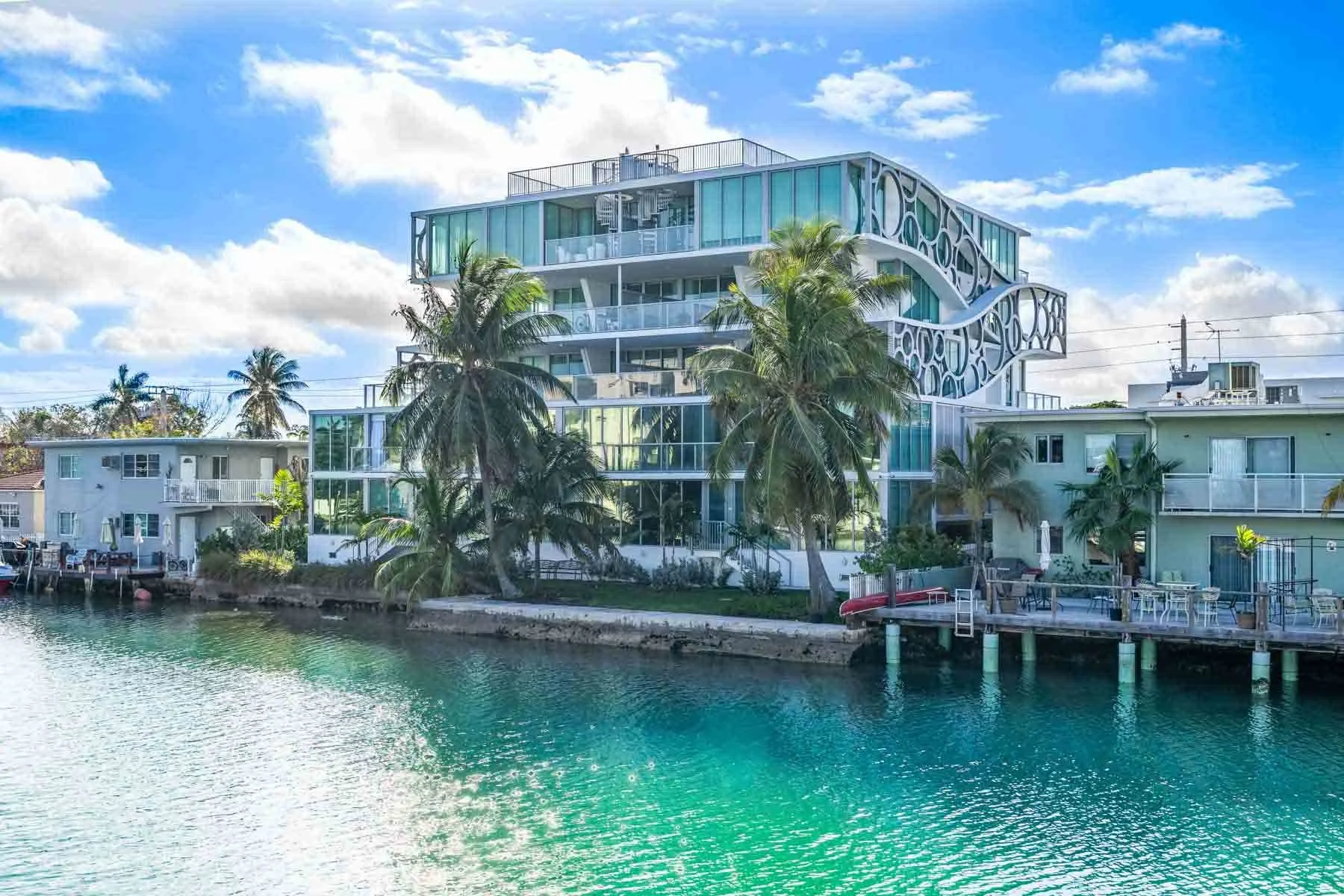 A waterfront scene with modern, multi-story buildings, palm trees, and a clear blue sky with scattered clouds.