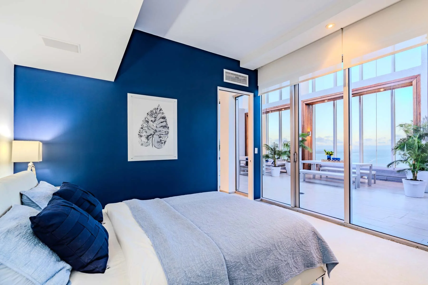 A bedroom with a blue accent wall, white bed, dark blue pillows, and artwork of a leaf, with a sliding glass door leading to an outdoor balcony with patio furniture and potted plants, overlooking the ocean.
