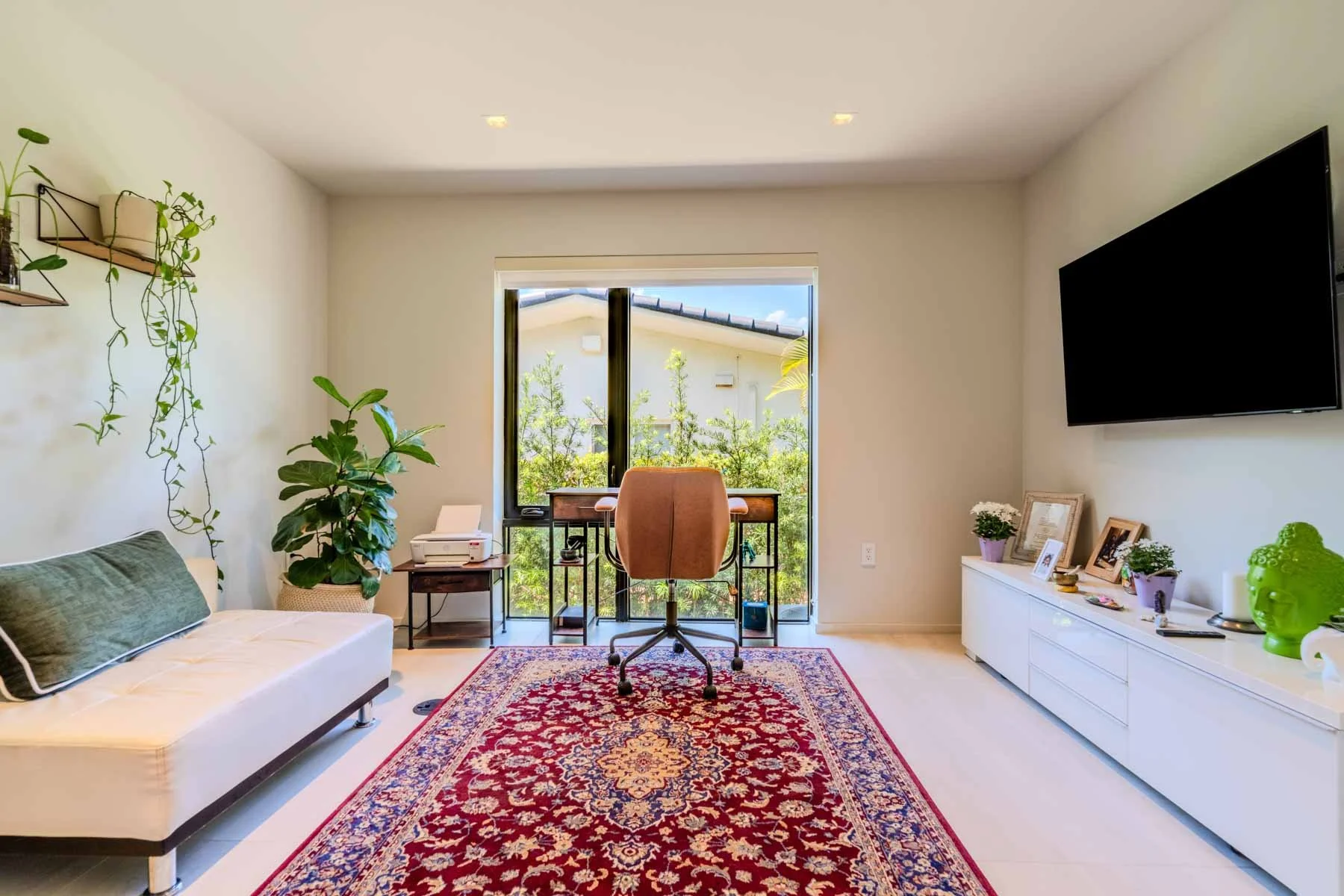 Living room with white walls, a window with greenery outside, a beige office chair at a desk, a white sofa with green pillows, potted plants, a colorful patterned rug, a large flat-screen TV, and framed photos on a white sideboard.