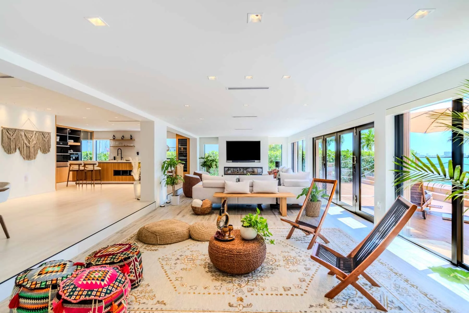 Bright, modern living room with white sofas, wooden chairs, and large sliding glass doors leading to an outdoor patio with bamboo plants.