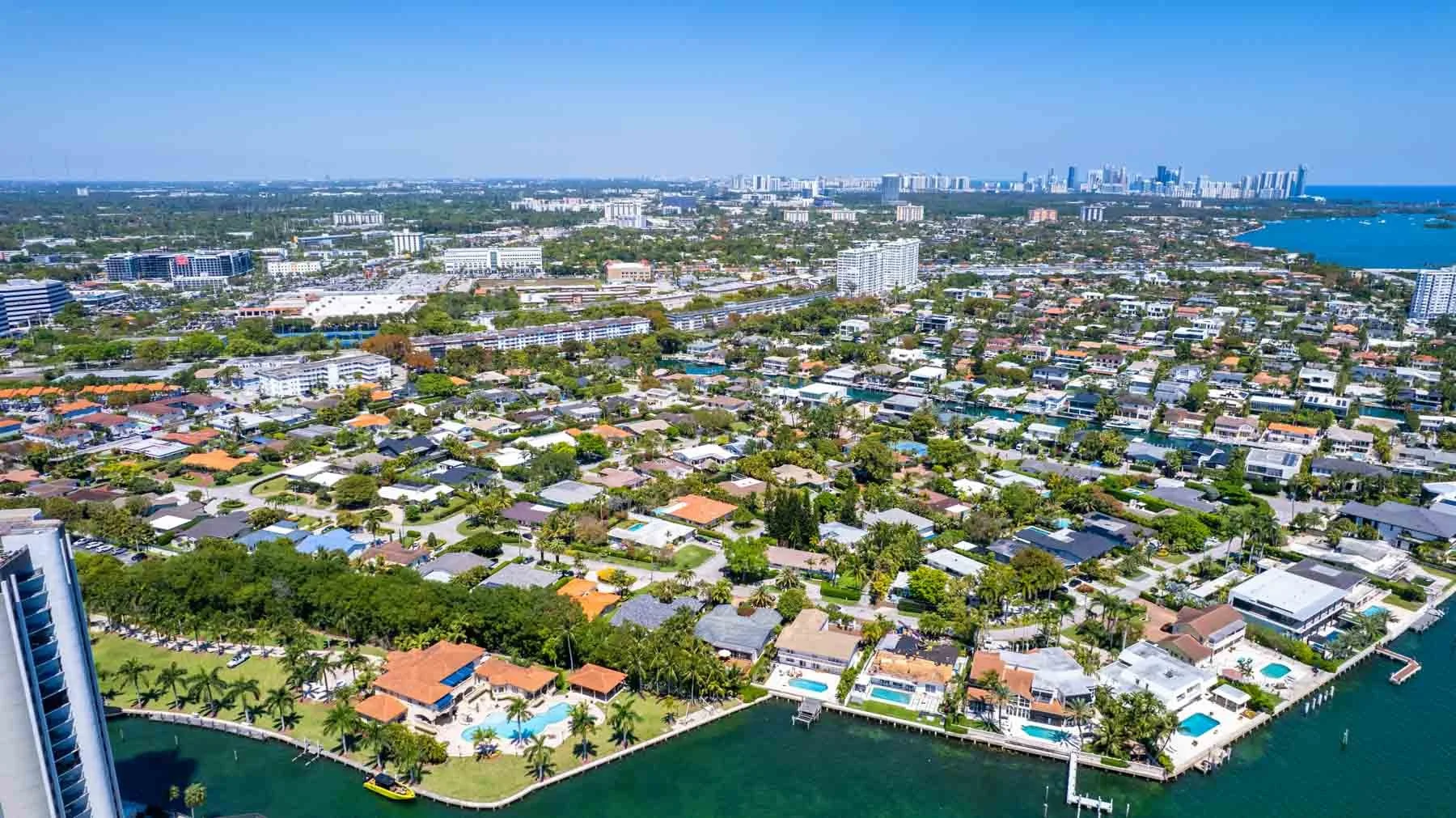 Aerial view of a coastal city with residential houses, high-rise buildings, and a harbor with boats, under a clear blue sky.