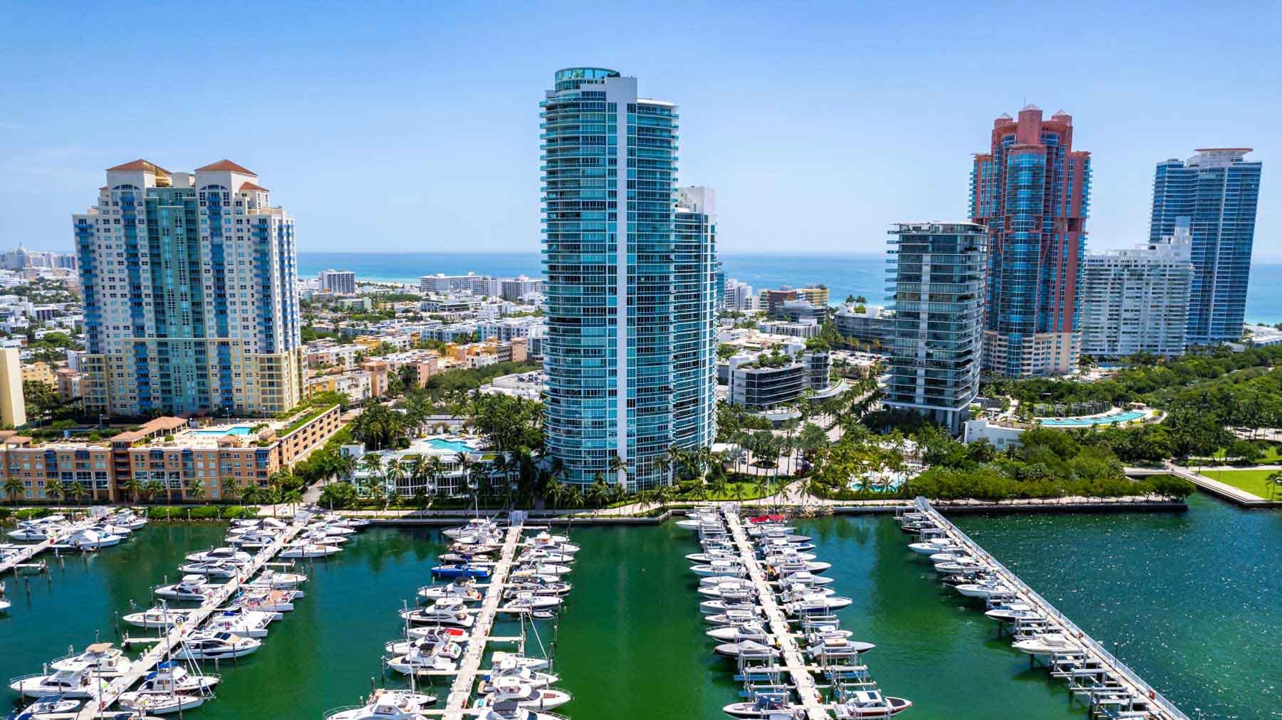 Aerial view of a marina with numerous yachts docked, tall modern skyscrapers overlooking the water, and the ocean in the background.
