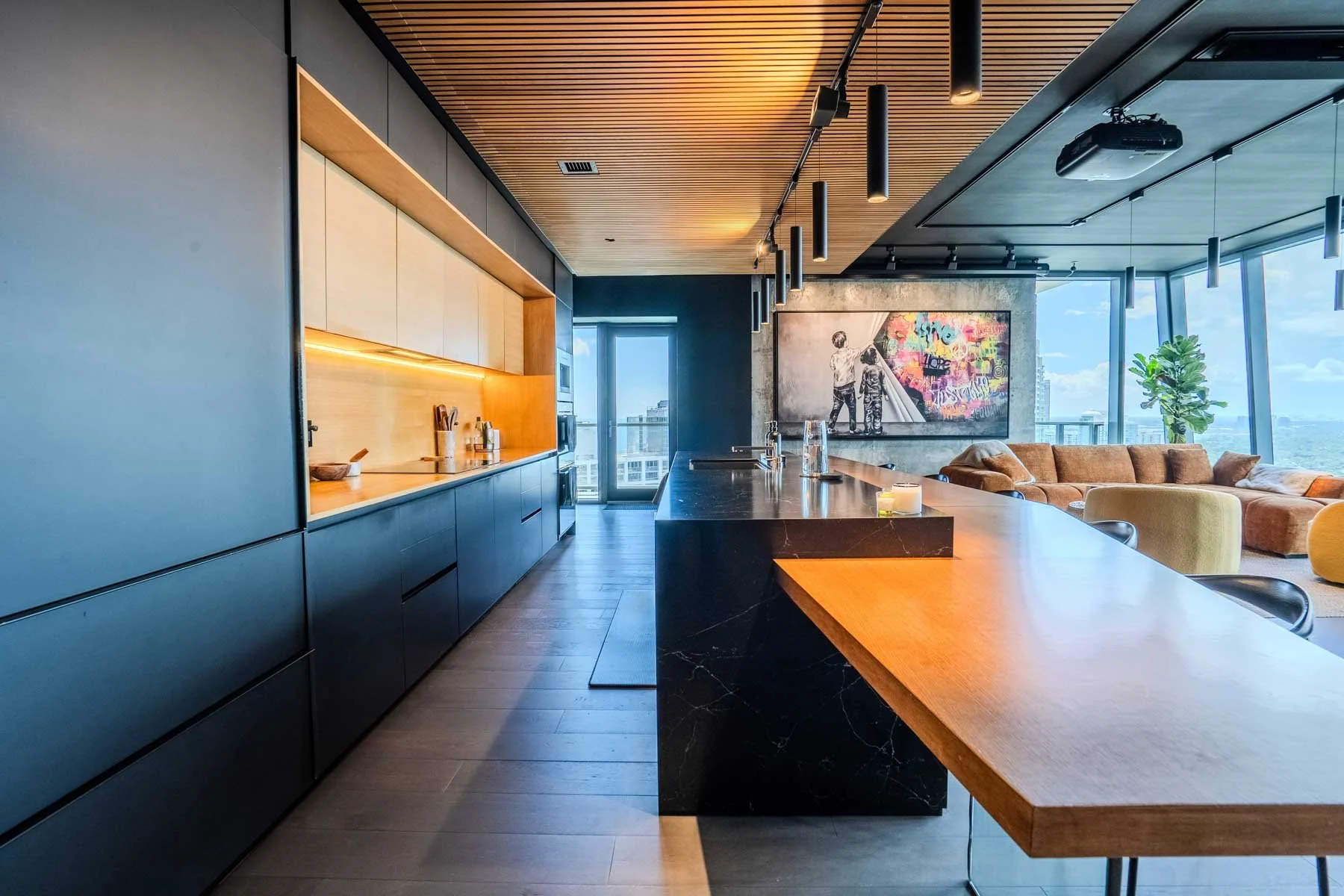 Modern open-concept kitchen and living room with a black marble kitchen island, wooden ceiling, large windows with city views, and contemporary furniture including a large sectional sofa.