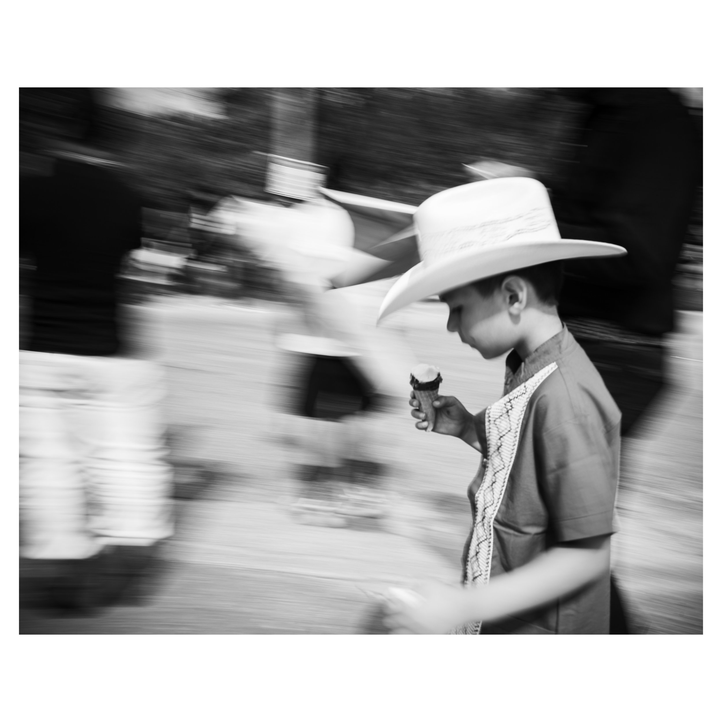 Street Photograph of a young boy in a cowboy hat eating an ice cream cone.  The photo is in black and white with a slow shutter drag efffect