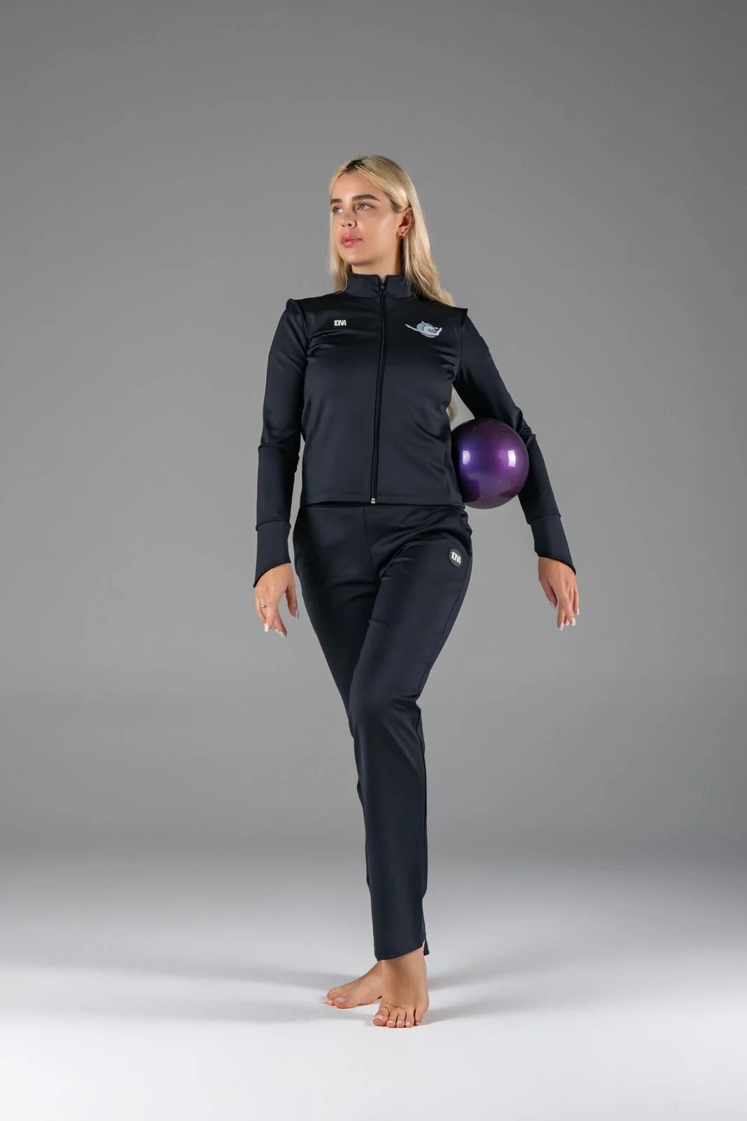 A woman standing on one foot holding a purple exercise ball, dressed in black athletic wear with a zipper jacket and matching pants, against a plain gray background.