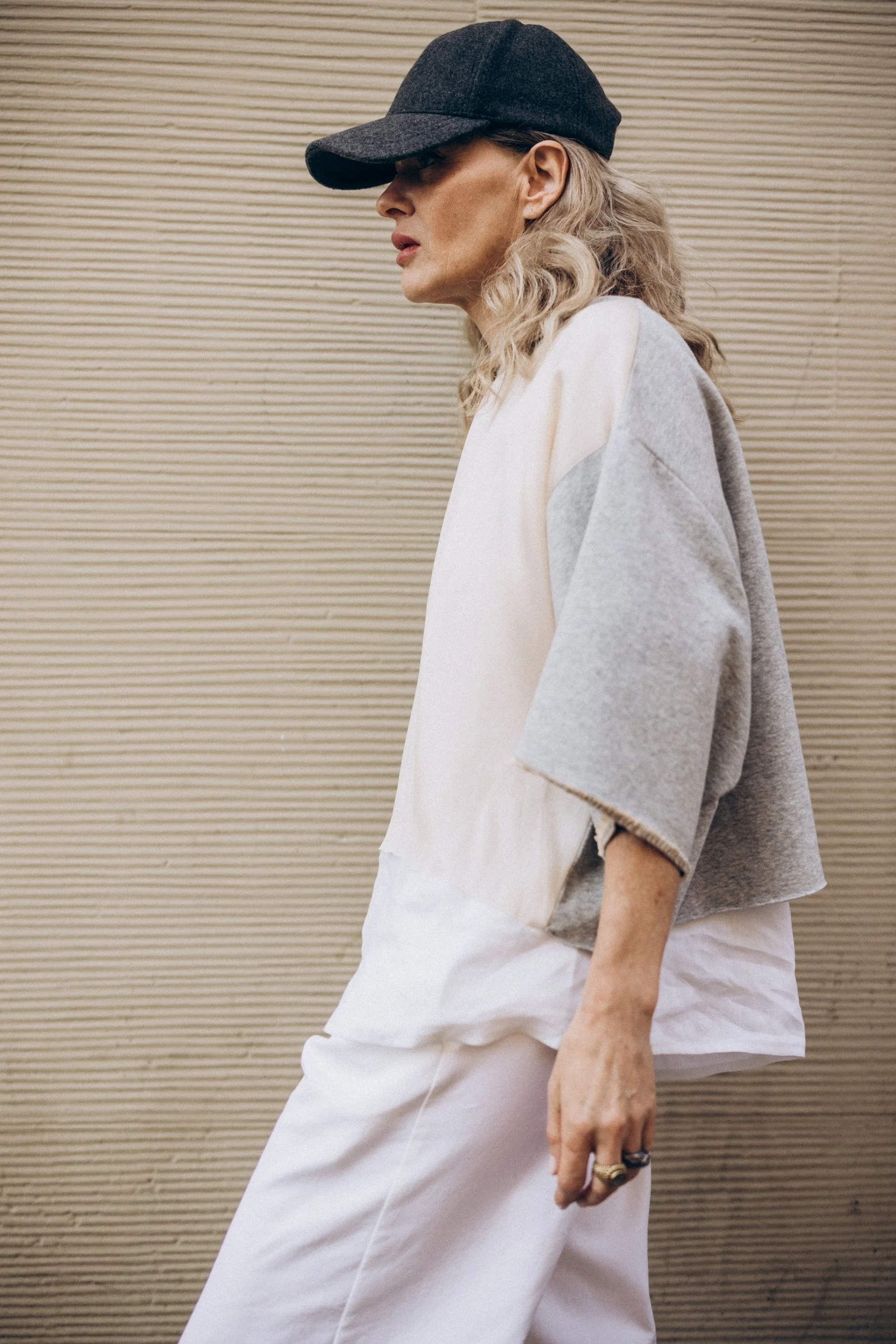 Side profile of a person with long blonde hair wearing a dark baseball cap, a loose-fitting color-blocked top, and white pants against a textured wall.