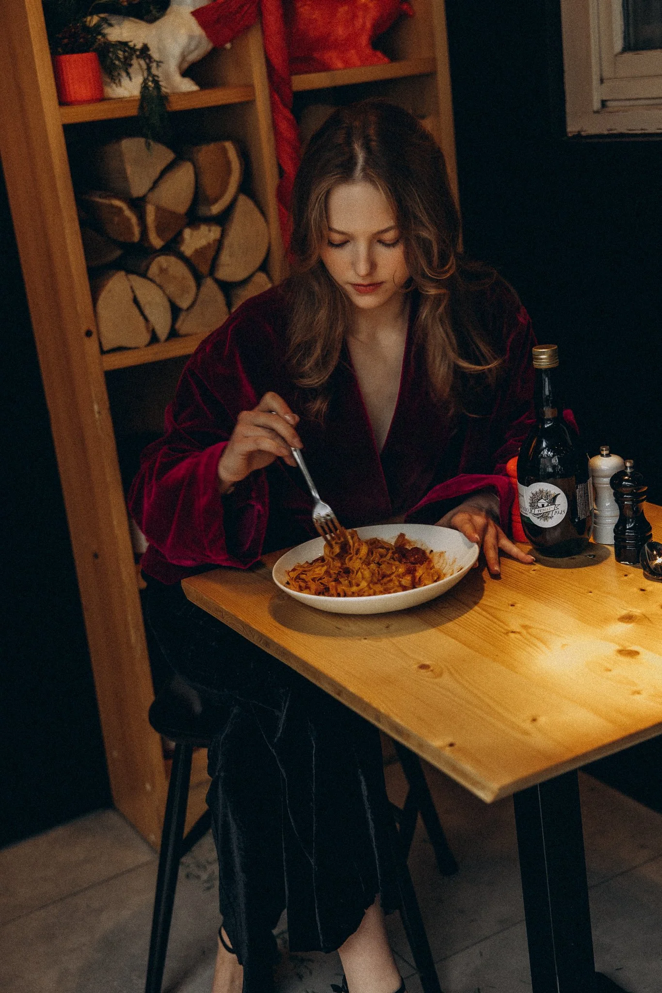 Woman in a red robe eating pasta at a wooden table with wine bottle nearby, shelves with logs in background.