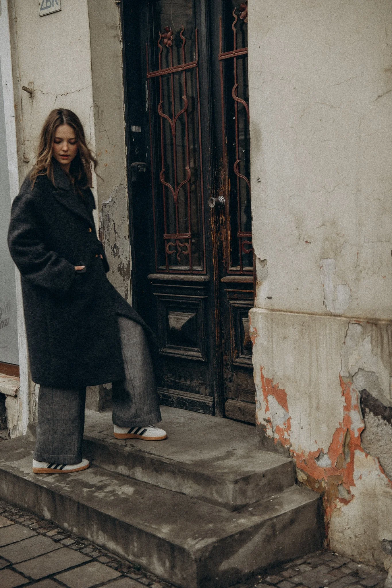 Woman in a dark coat standing on concrete steps in front of an old, worn door with decorative metalwork, beside a weathered wall.