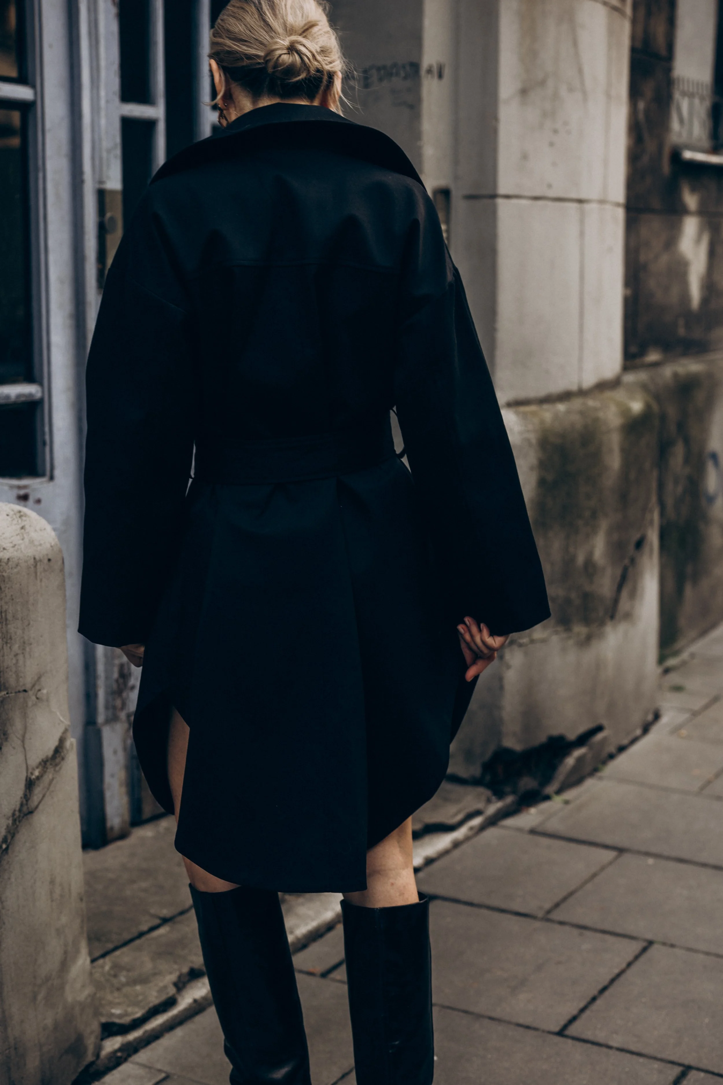 Person wearing a long black coat and boots walking on a city sidewalk
