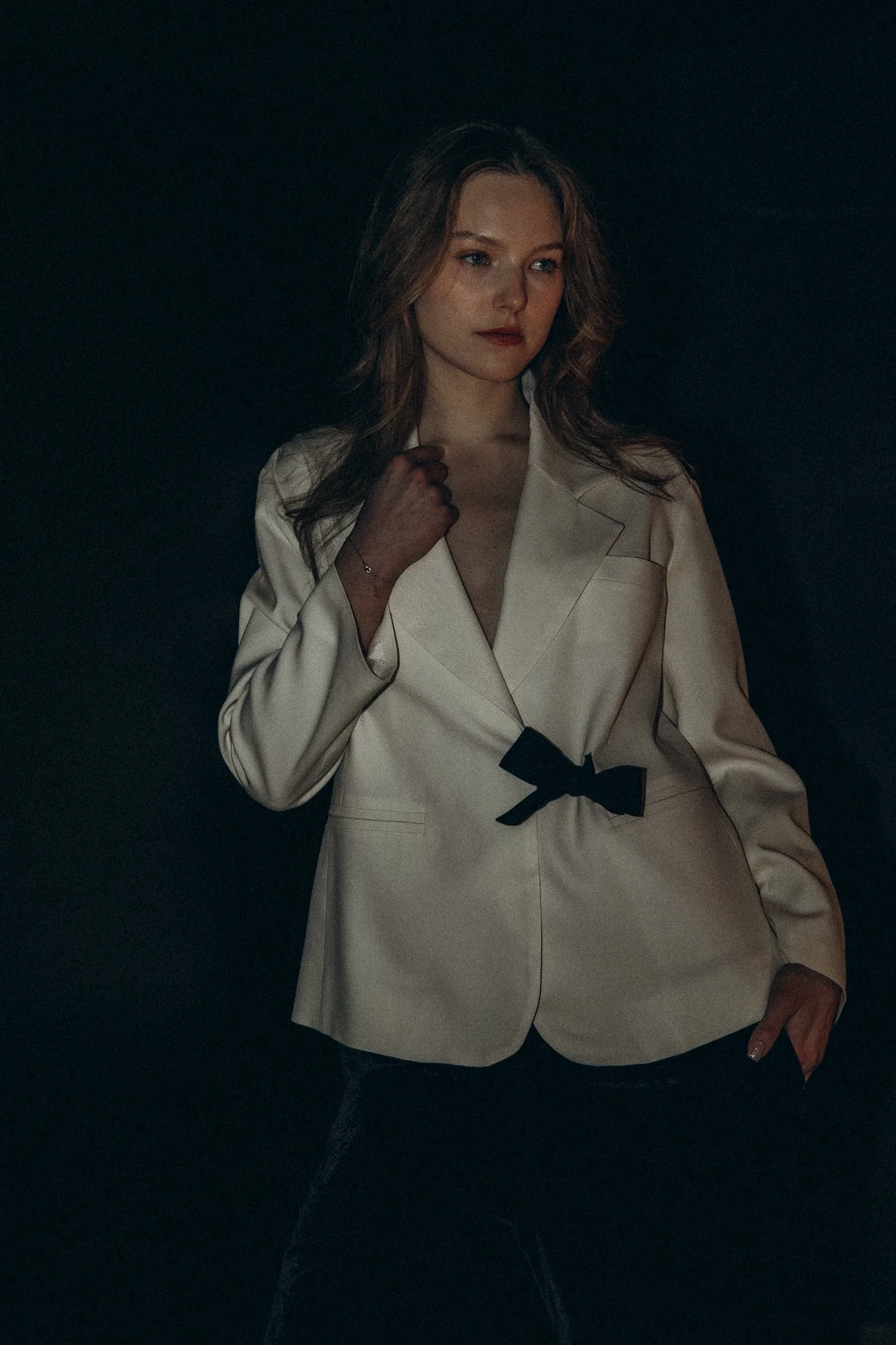 Woman wearing a white blazer with black bow detail, standing against a dark background.