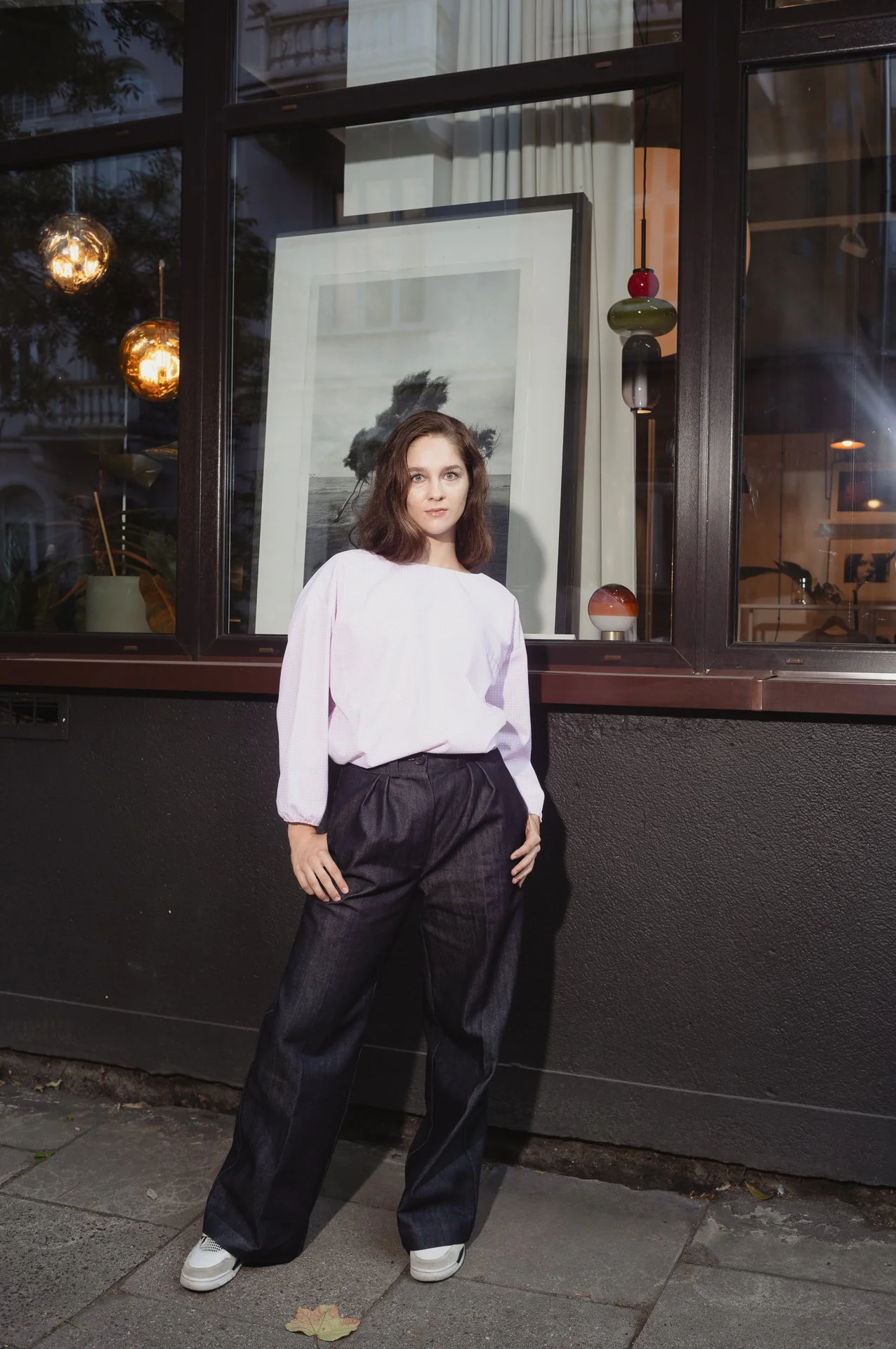 Person wearing a white blouse and dark jeans stands in front of a window with art inside, next to hanging lights.
