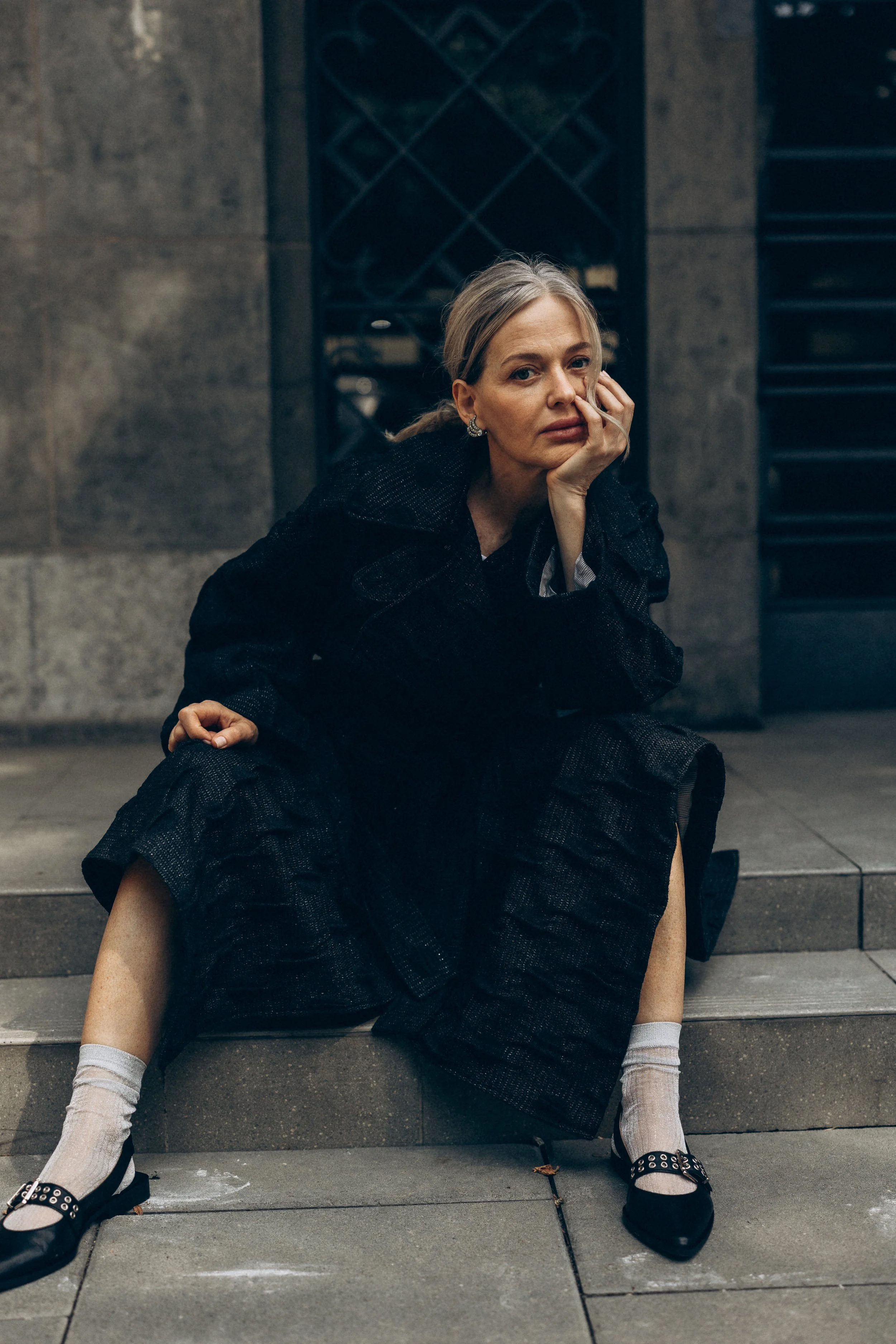 A woman sitting on outdoor steps wearing a dark coat, light gray socks, and black shoes, with her head resting on her hand.