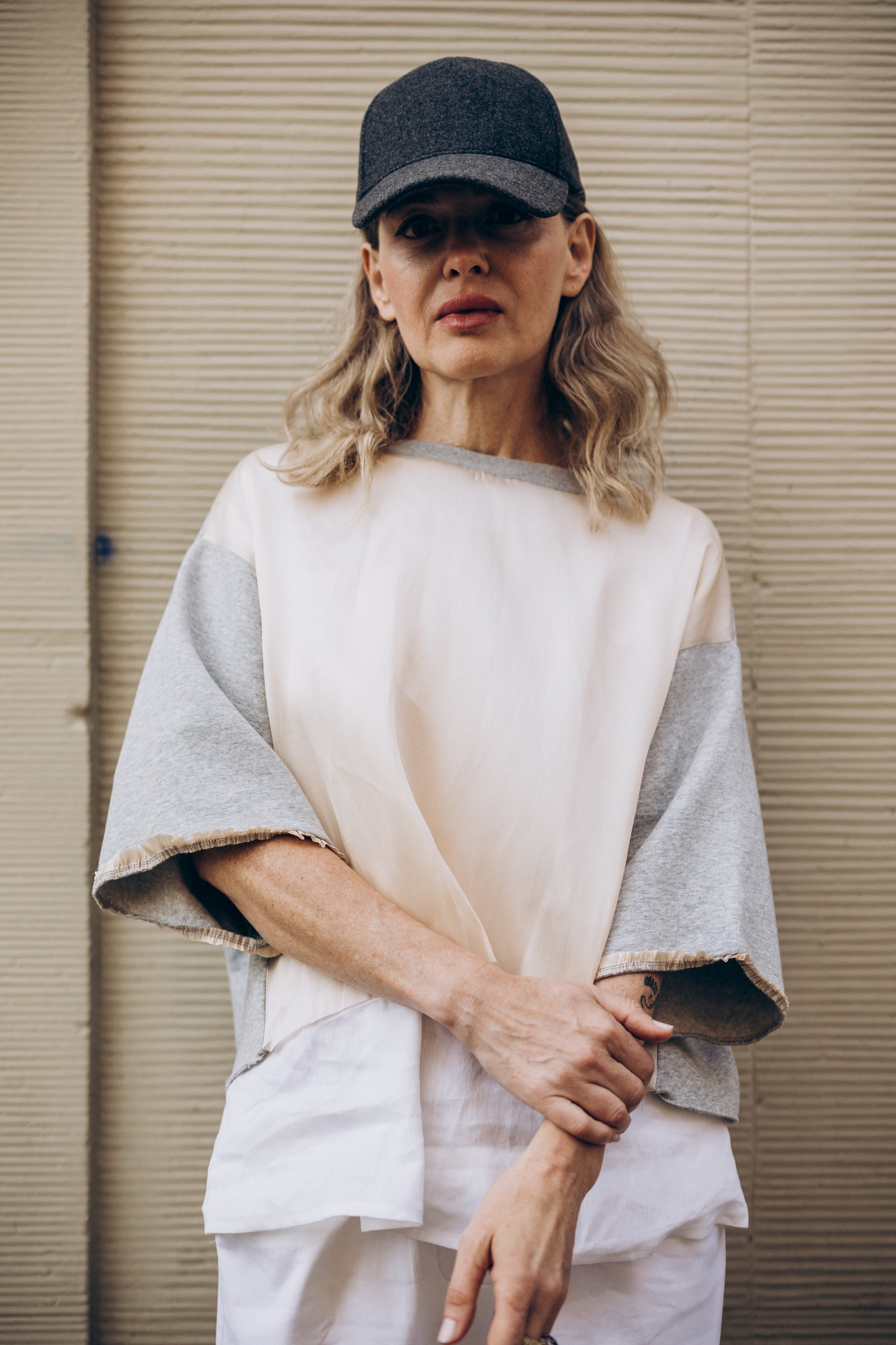 Woman wearing a baseball cap and casual clothing standing against a textured wall.