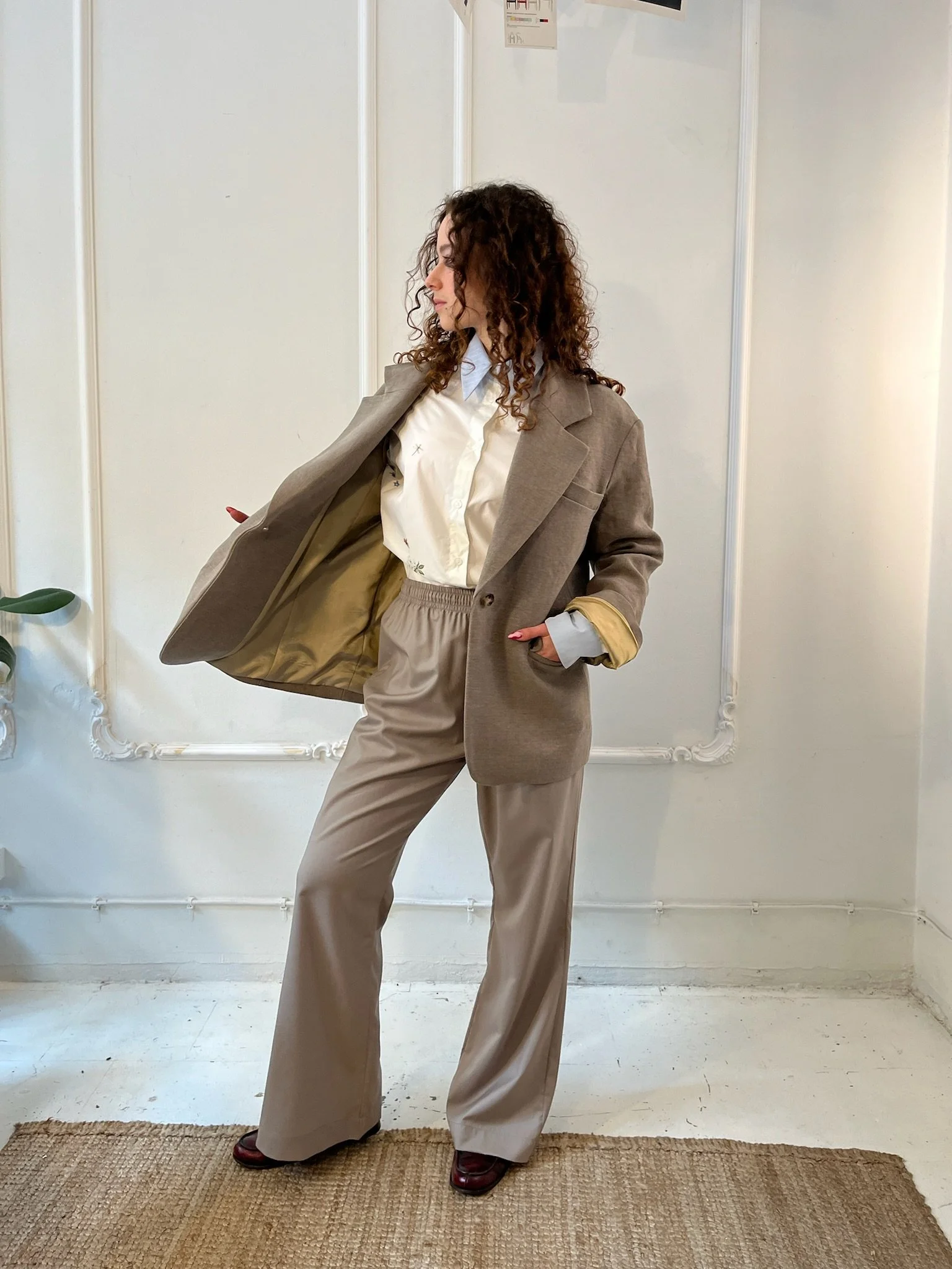 Woman wearing a beige suit and white shirt, posing indoors against a white wall.