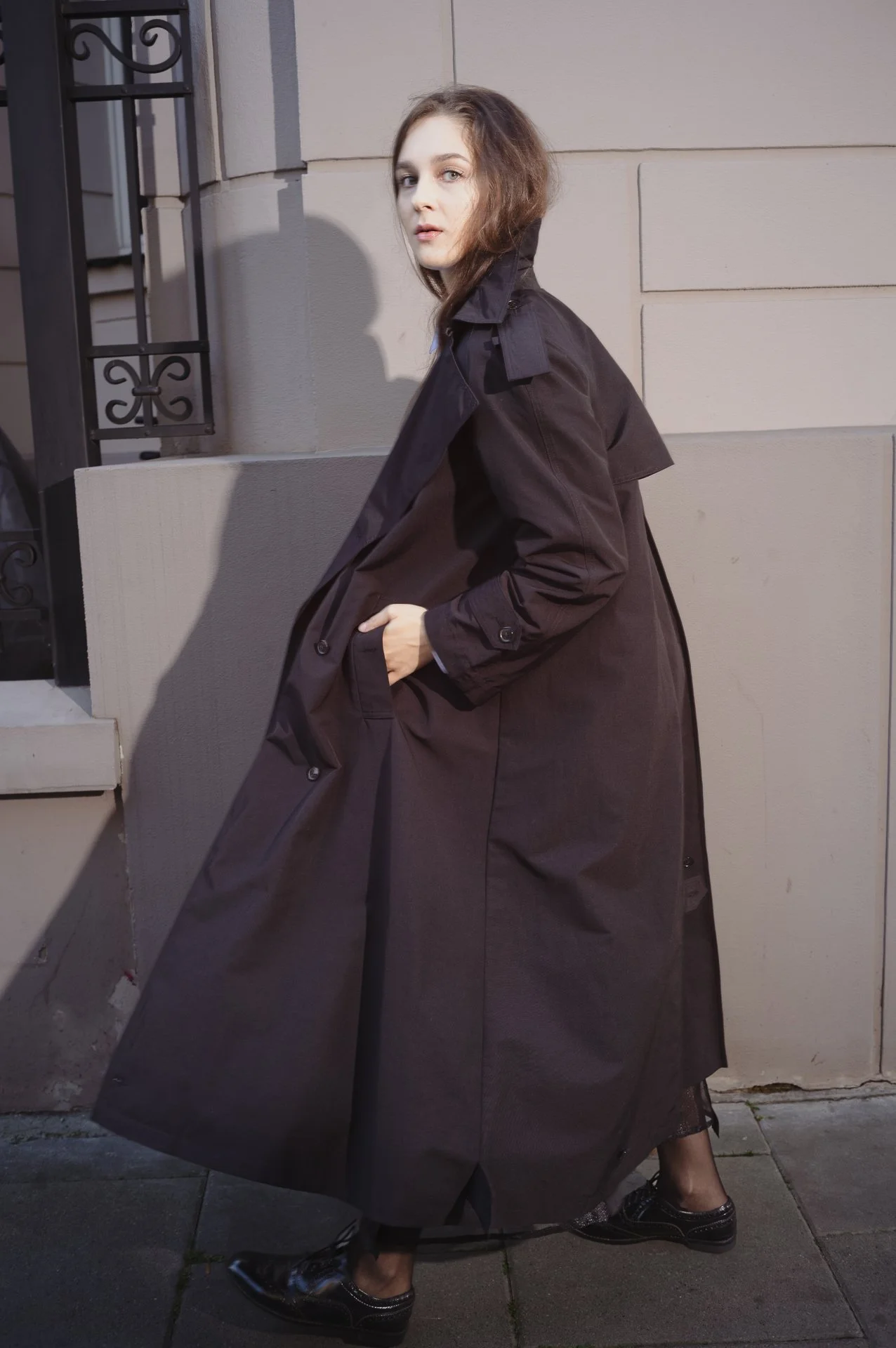 Person wearing a long dark trench coat standing on a sidewalk.