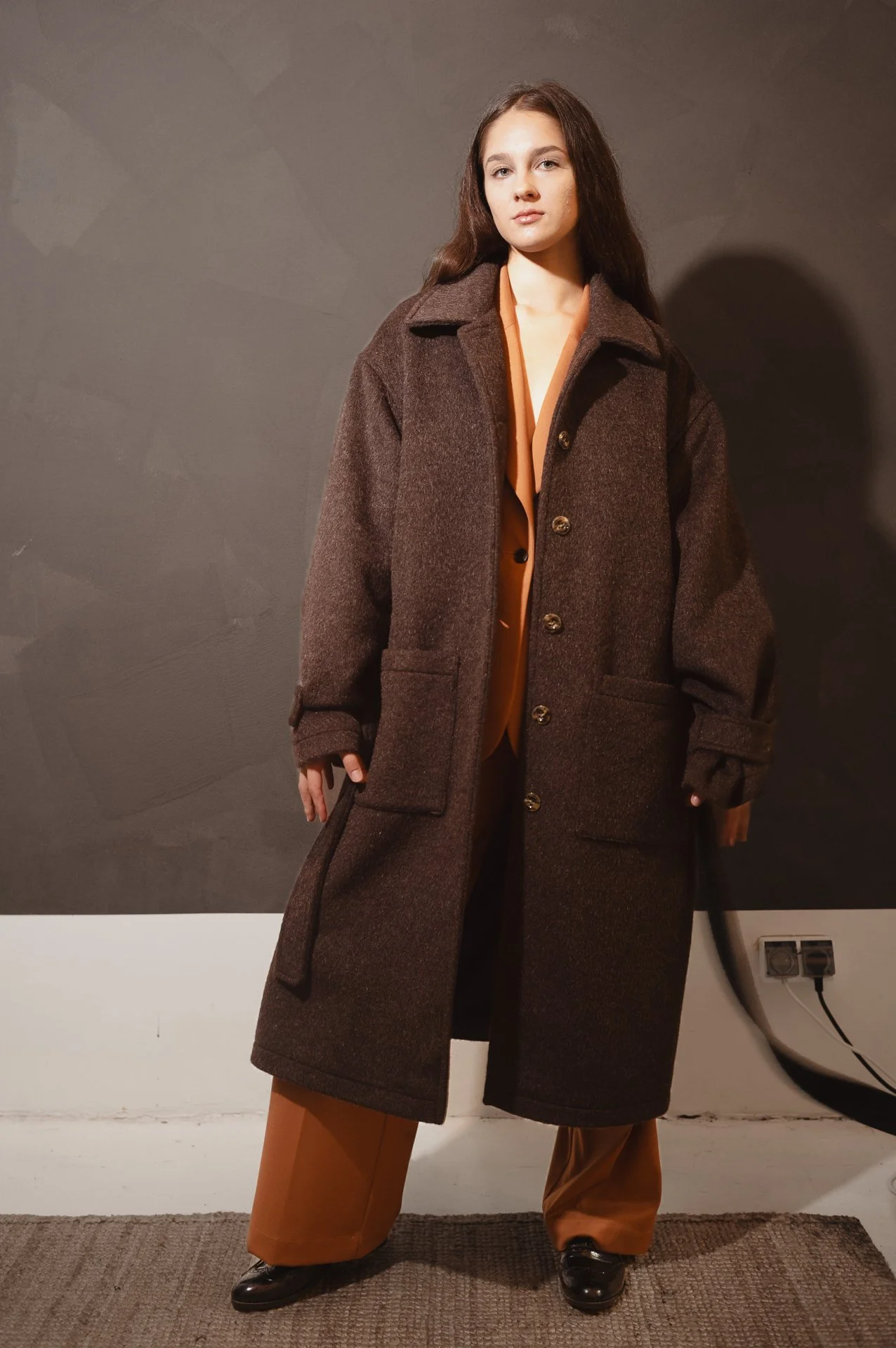 Woman wearing a long brown overcoat over a tan outfit, standing against a neutral background indoors.