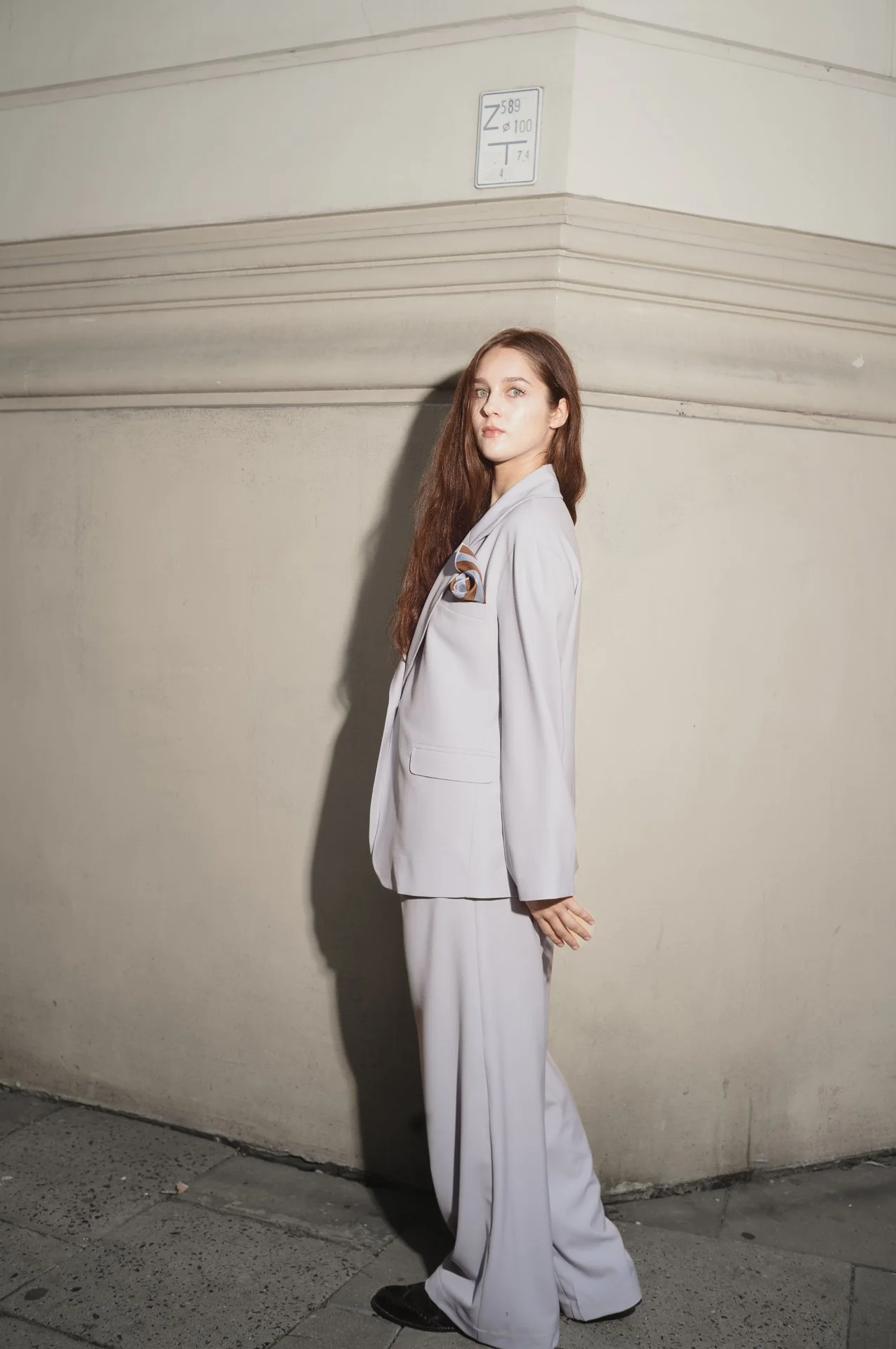 Person wearing a light gray suit standing against a beige wall outdoors.