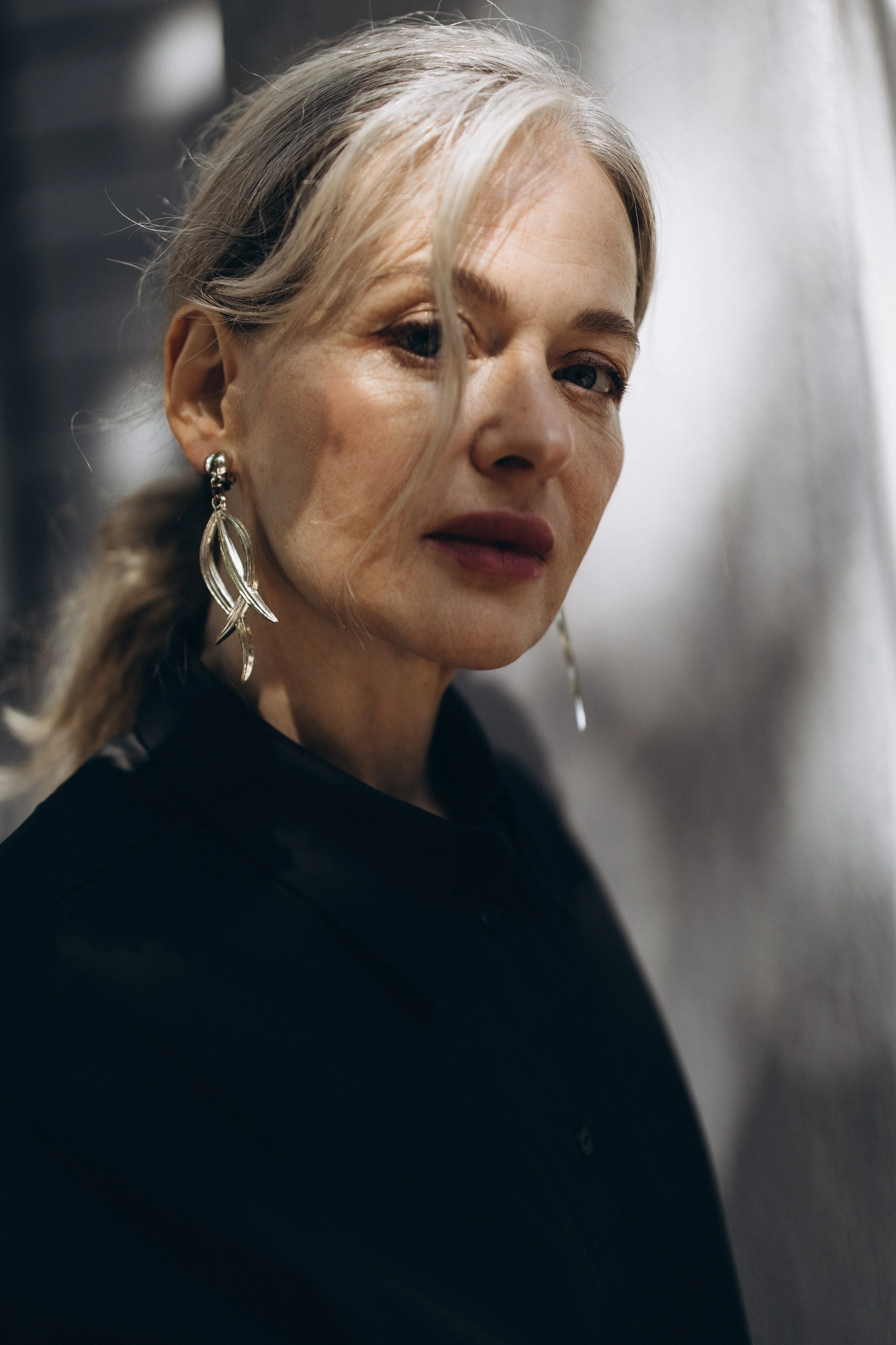 Portrait of a woman with light gray hair and long earrings, wearing a dark shirt, looking at the camera.