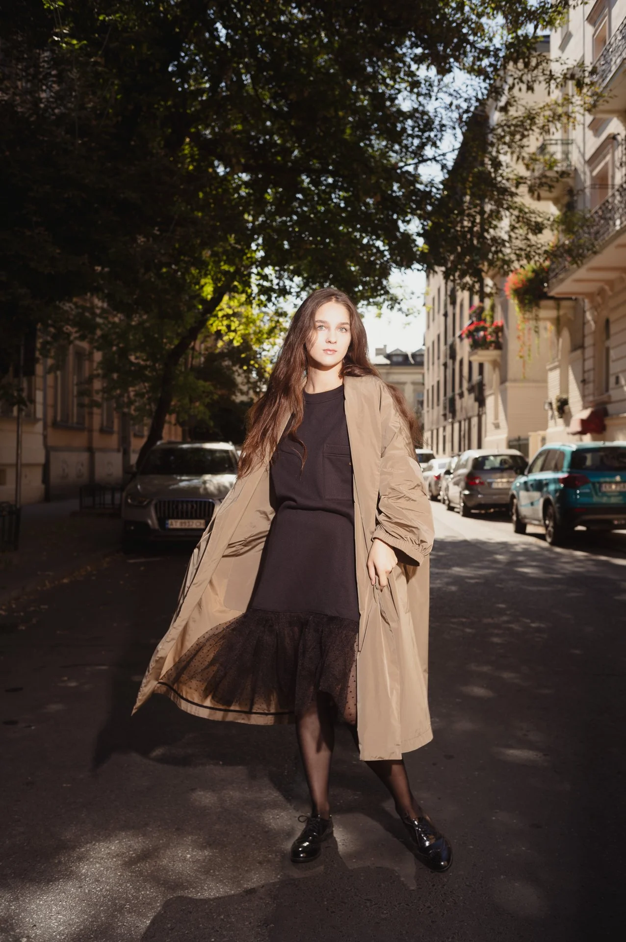 Woman wearing a tan coat over a black dress stands on a sunlit street with trees and parked cars in the background.