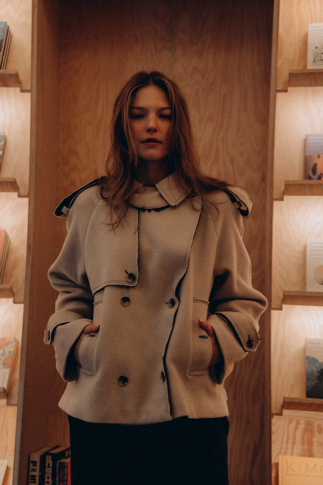 Person in a beige coat standing in front of bookshelves.