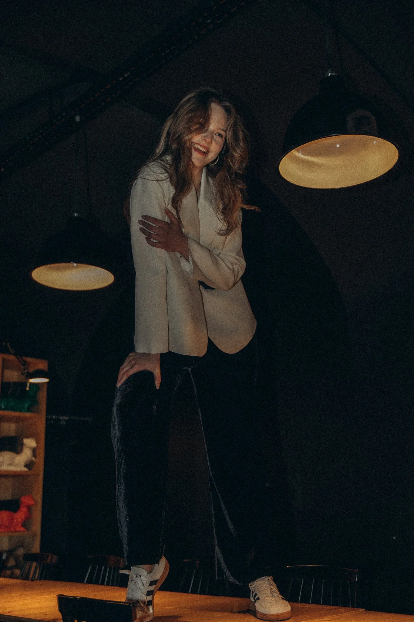 Person wearing a white blazer and black pants standing on a table in a dimly lit room.
