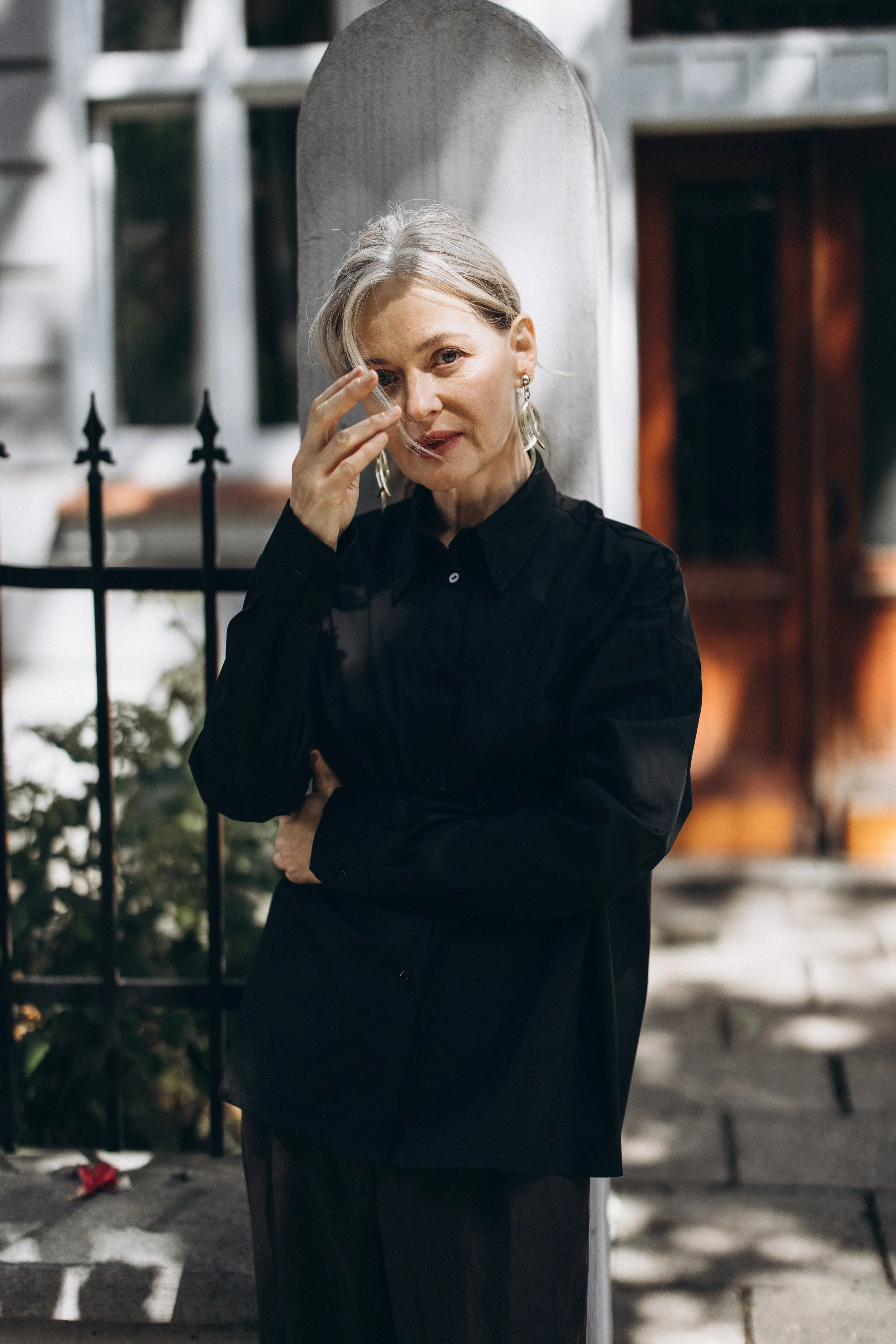 Woman with gray hair in black shirt standing outdoors, holding her hair, with a fence and building in the background.