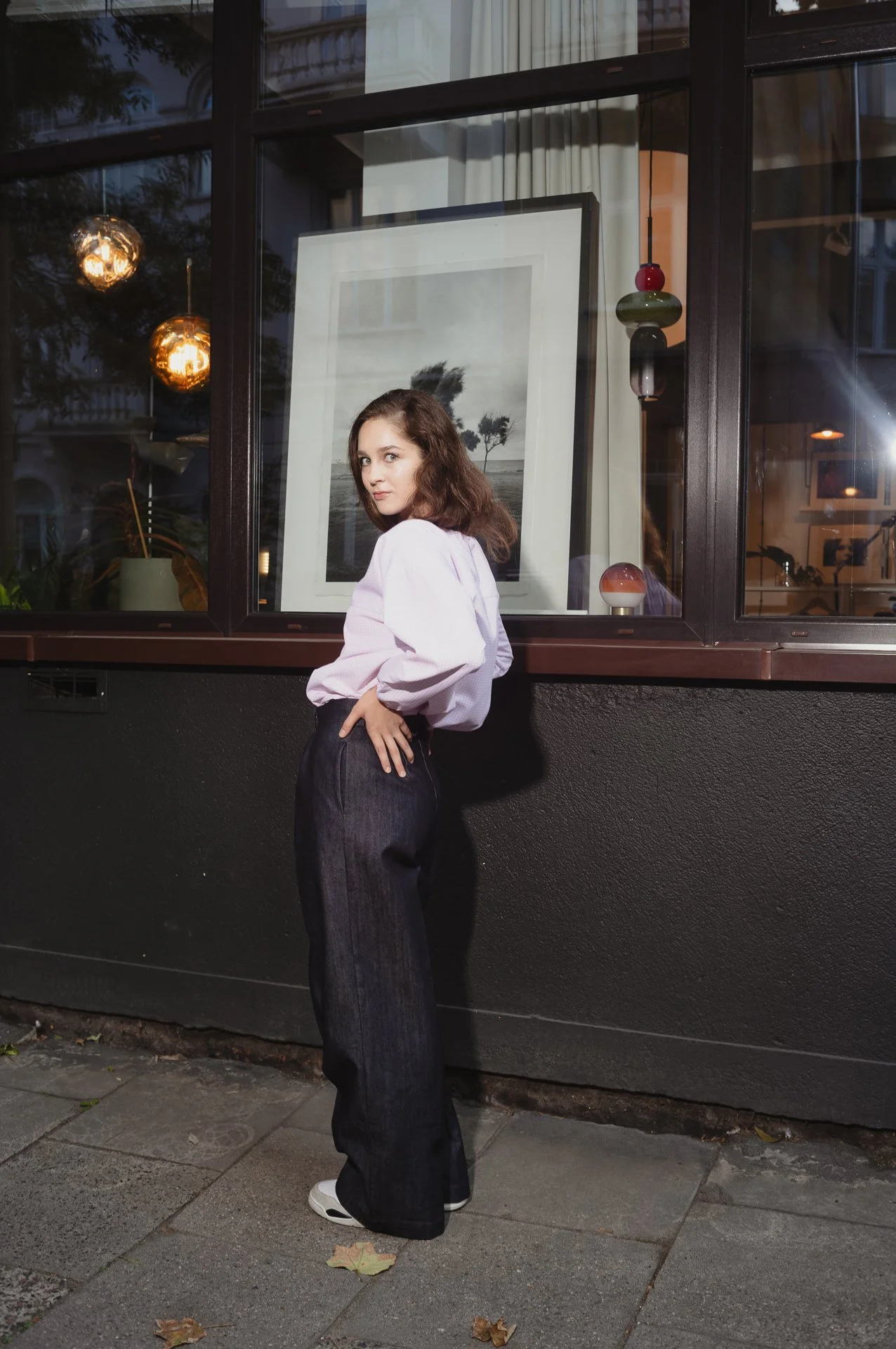 Woman posing in front of a window wearing a light-colored top and dark pants, with reflections and hanging lights in the background.