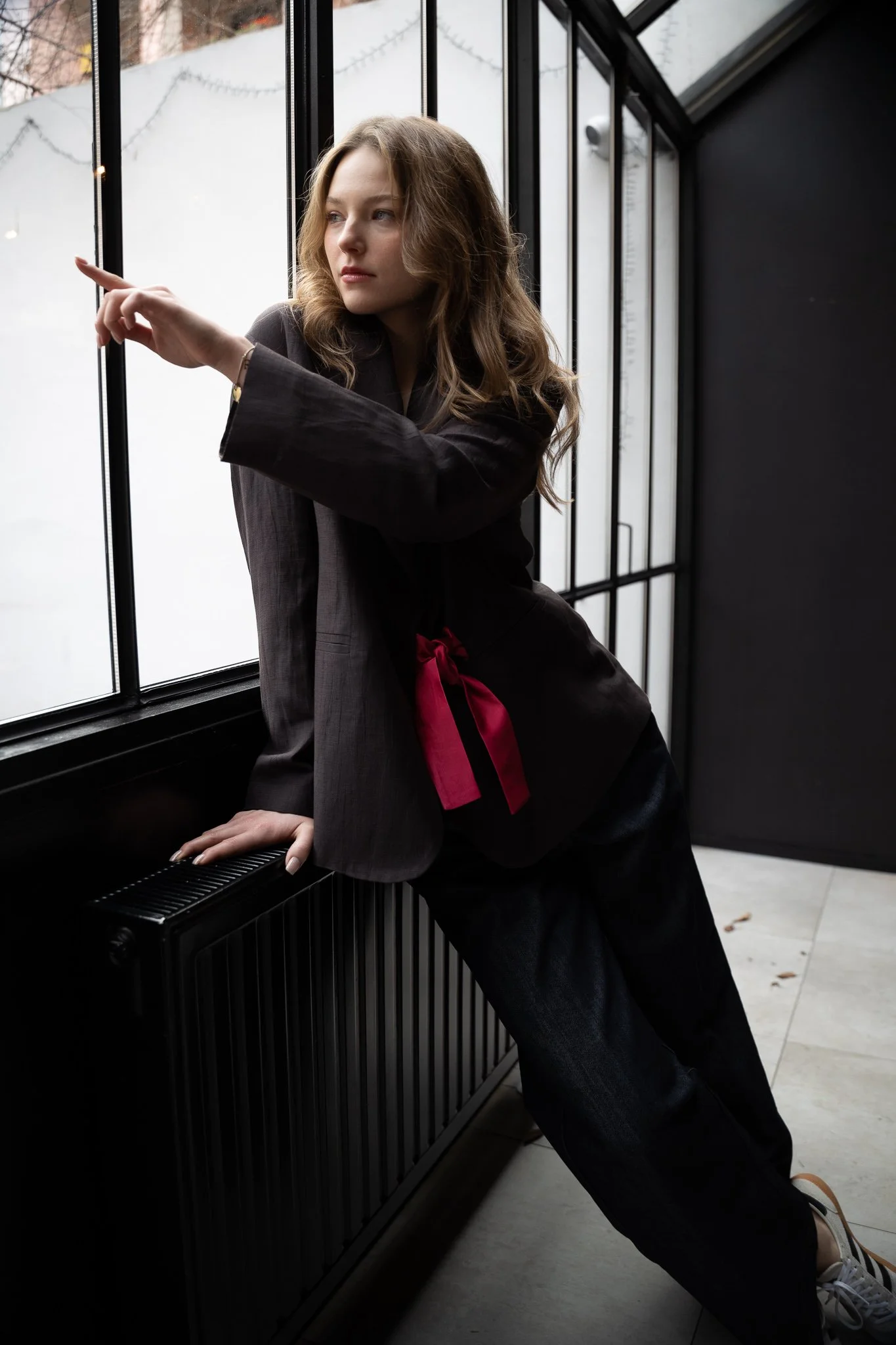 Woman in dark blazer with red ribbon leaning against a window, pointing outside.