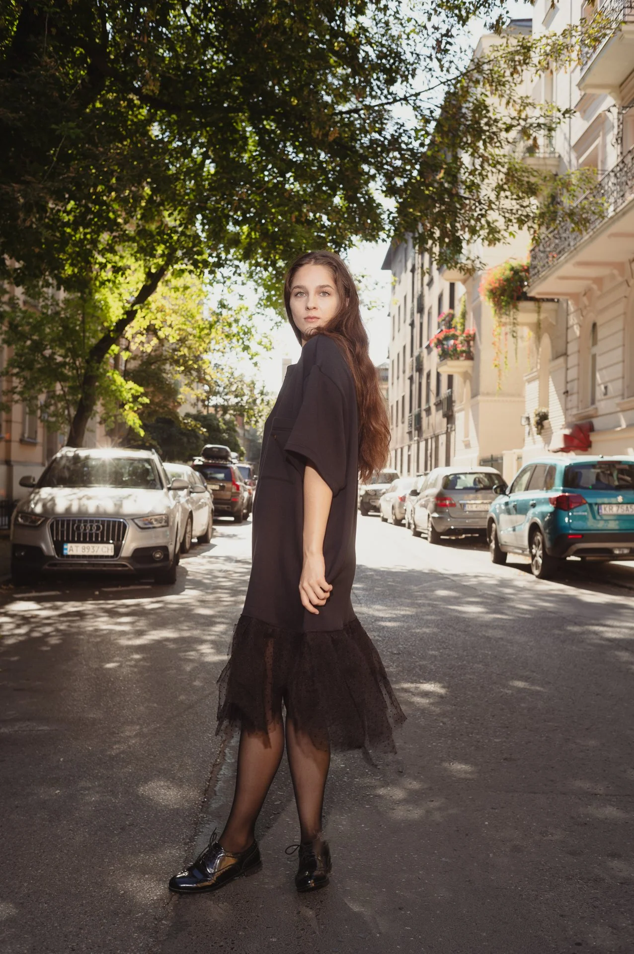 Woman in black dress standing on a tree-lined street with cars parked on both sides.