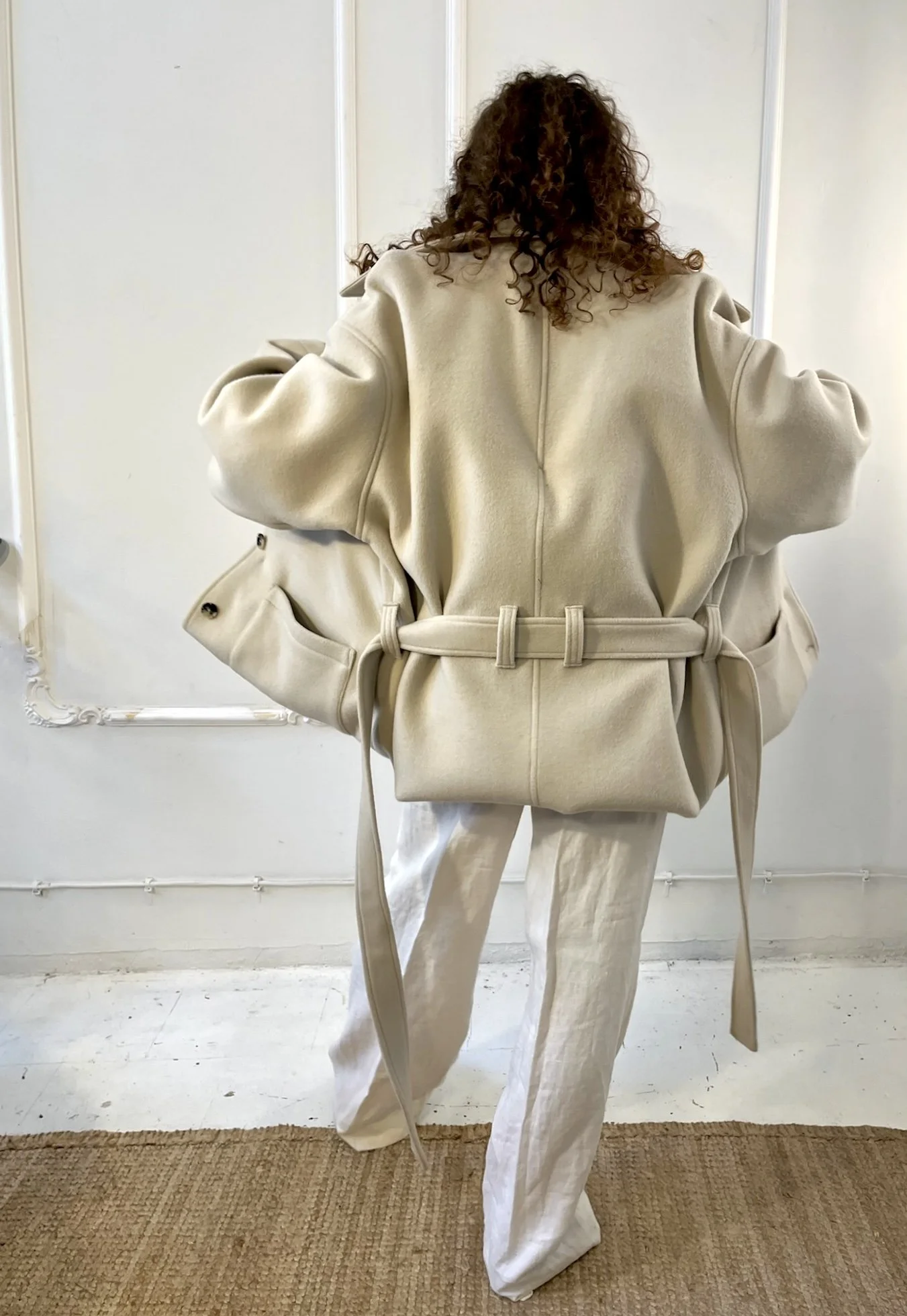 Person wearing oversized beige coat with tied belt and white pants, standing on a woven rug in a white room.