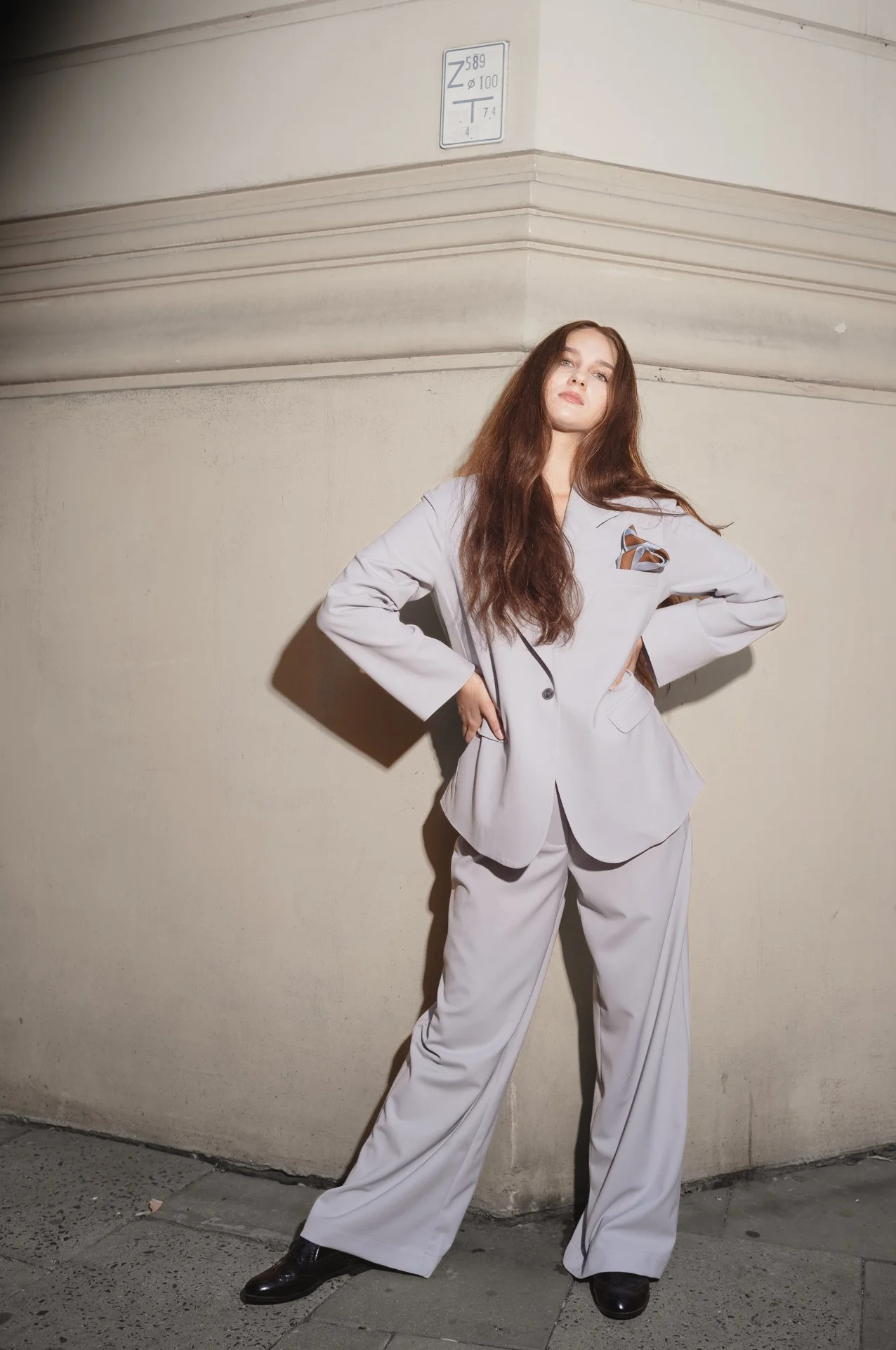 Person wearing a light gray suit standing against a beige wall with hands on hips, street scene.