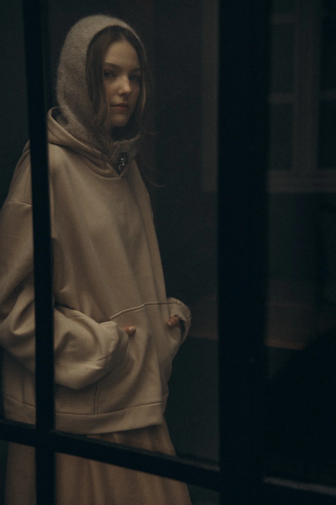 Young woman in a hooded sweater, seen through a window, with a neutral expression.