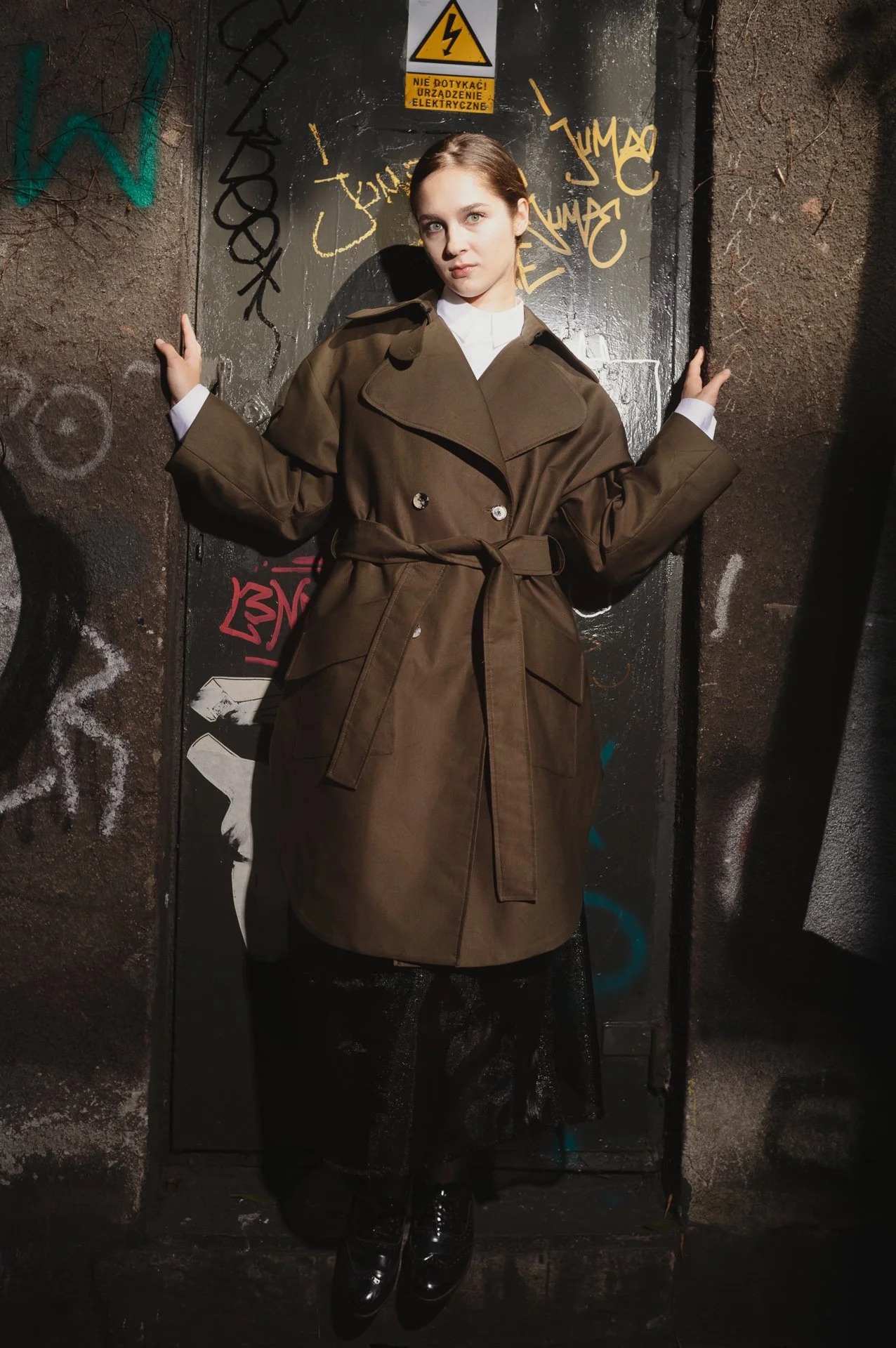 Person wearing olive trench coat standing against a graffiti-covered wall.
